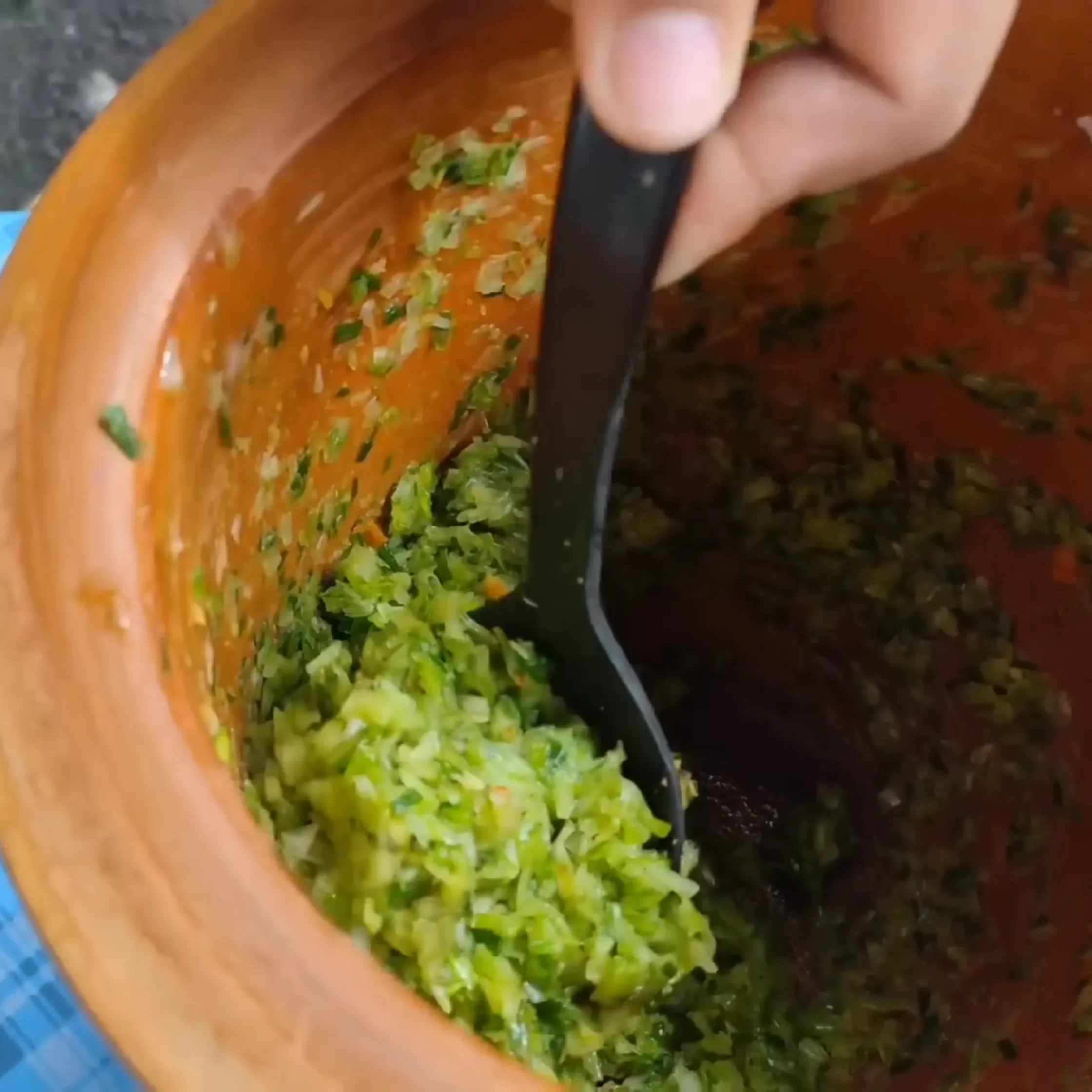 Close-up of chopped green herbs stirred into an orange sauce in a wooden bowl with a black spatula visible in the foreground.