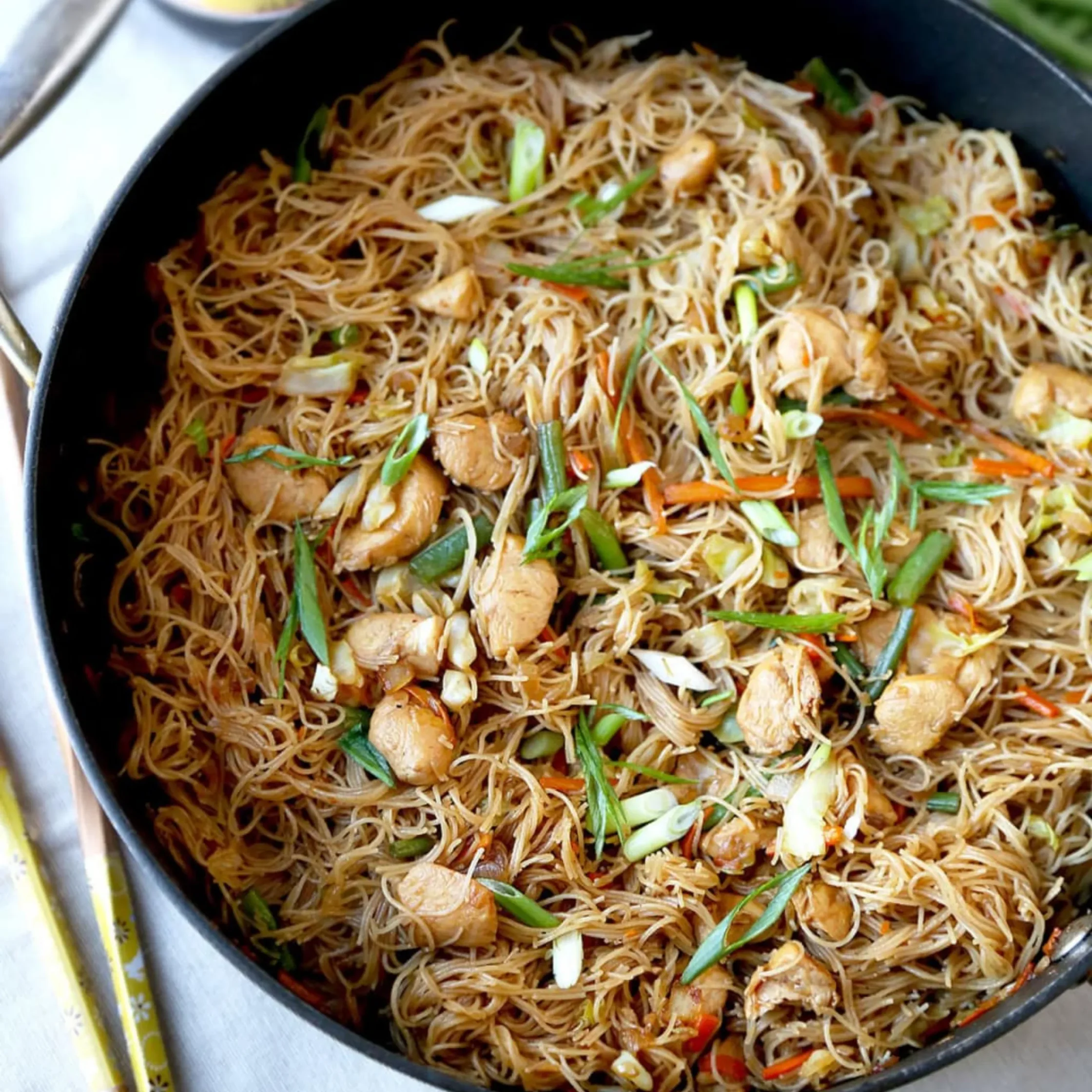 Black skillet filled with stir-fried rice noodles, chicken, vegetables, and herbs, with chopsticks and condiment bottles nearby; appetizing, casual meal.