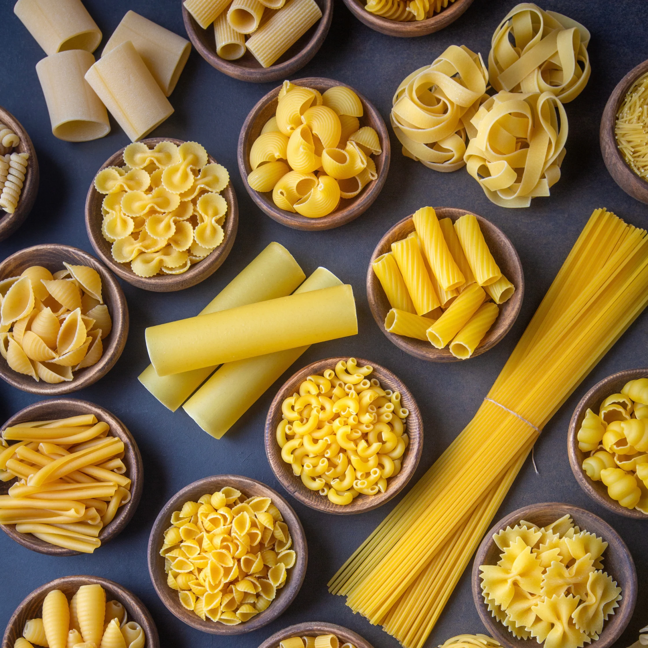 Top-down still life of assorted dry pasta—rigatoni, shells, fusilli, farfalle, macaroni, tagliatelle, and spaghetti—in bowls on dark slate.