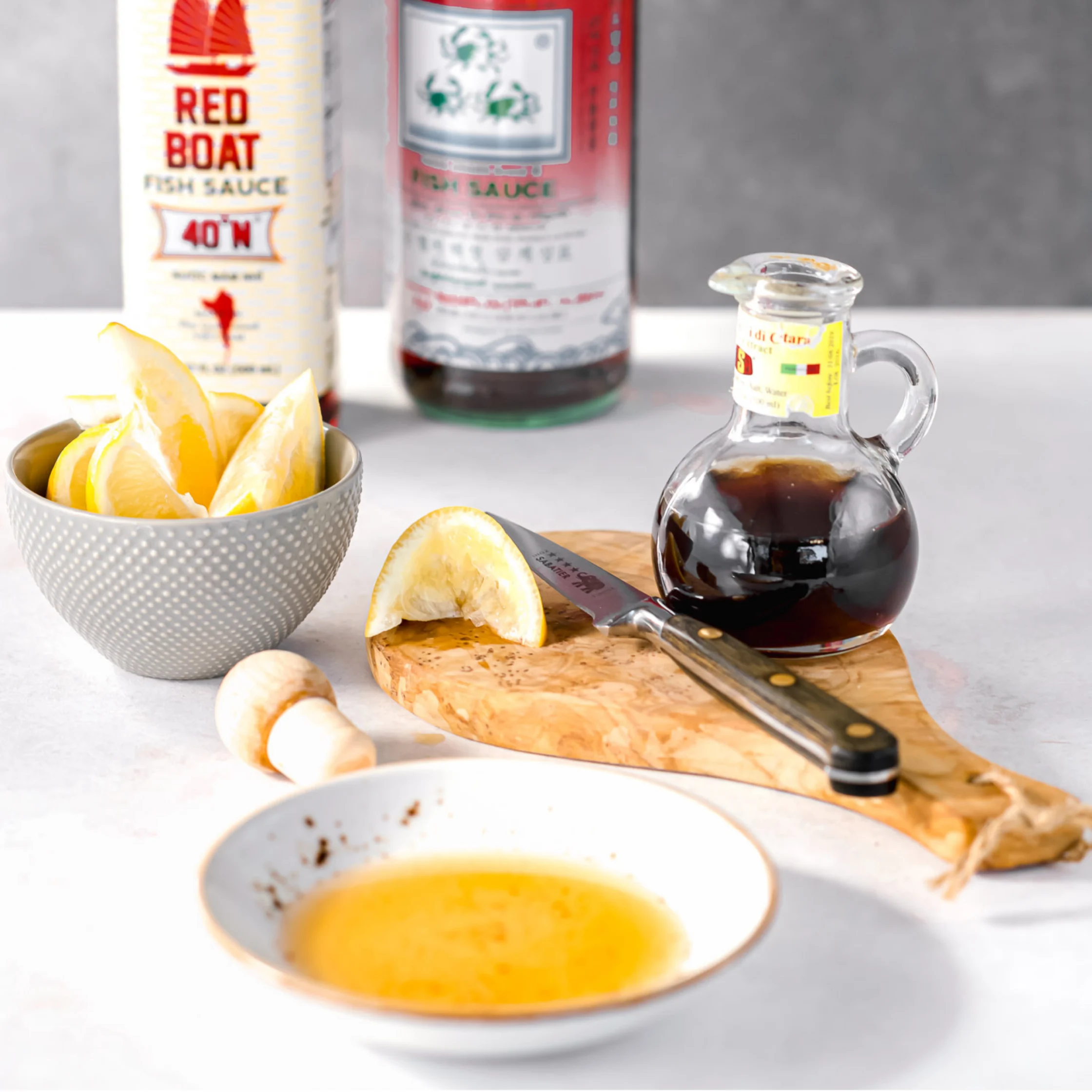 A wooden board holds a halved lemon and knife beside bowls of lemon wedges and golden dipping sauce, with a glass jug of dark soy sauce and Red Boat fish sauce behind. The bright kitchen still life feels fresh and inviting.