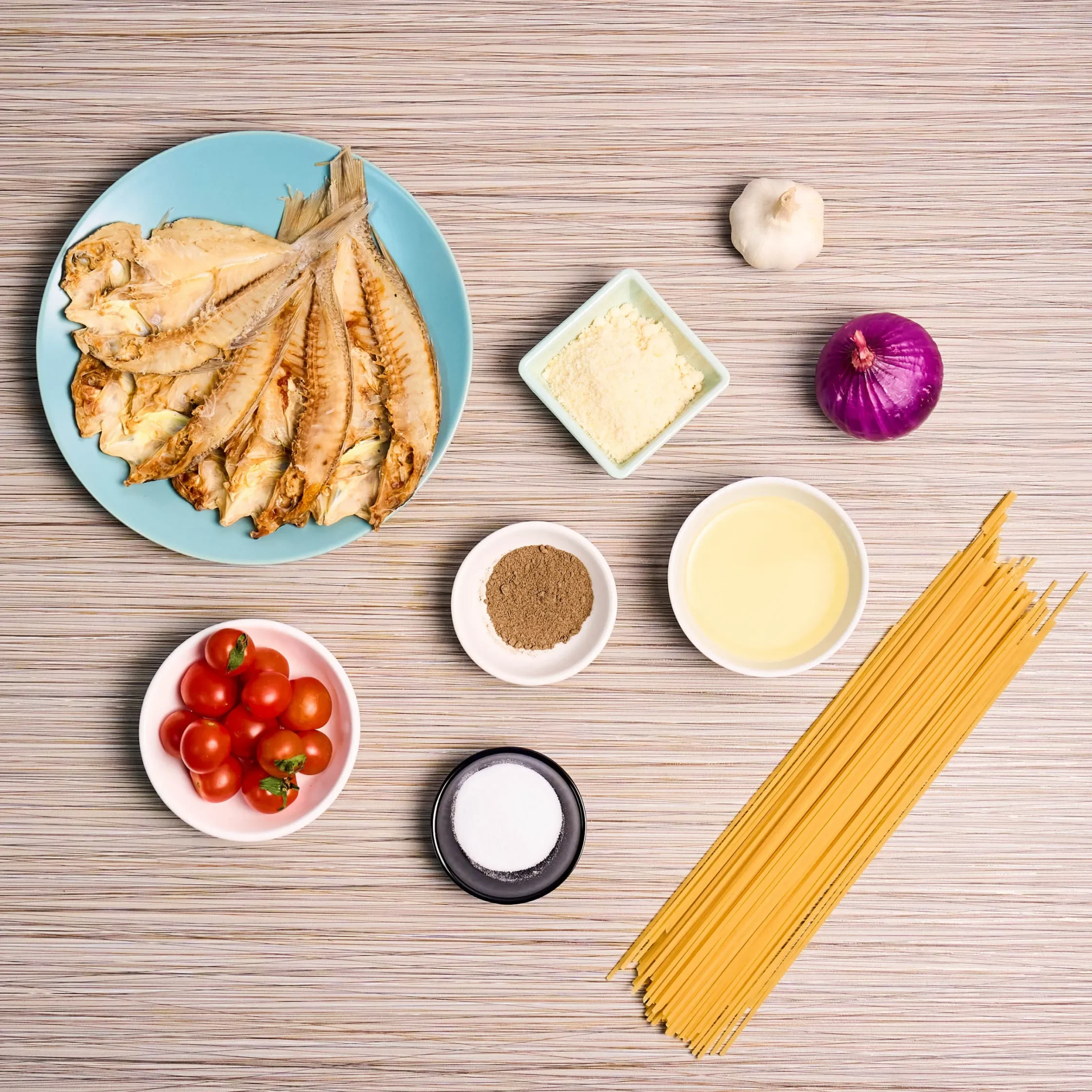 Plate of dried fish fillets on a light blue plate with garlic, flour, tomatoes, and uncooked spaghetti nearby on a wooden surface.