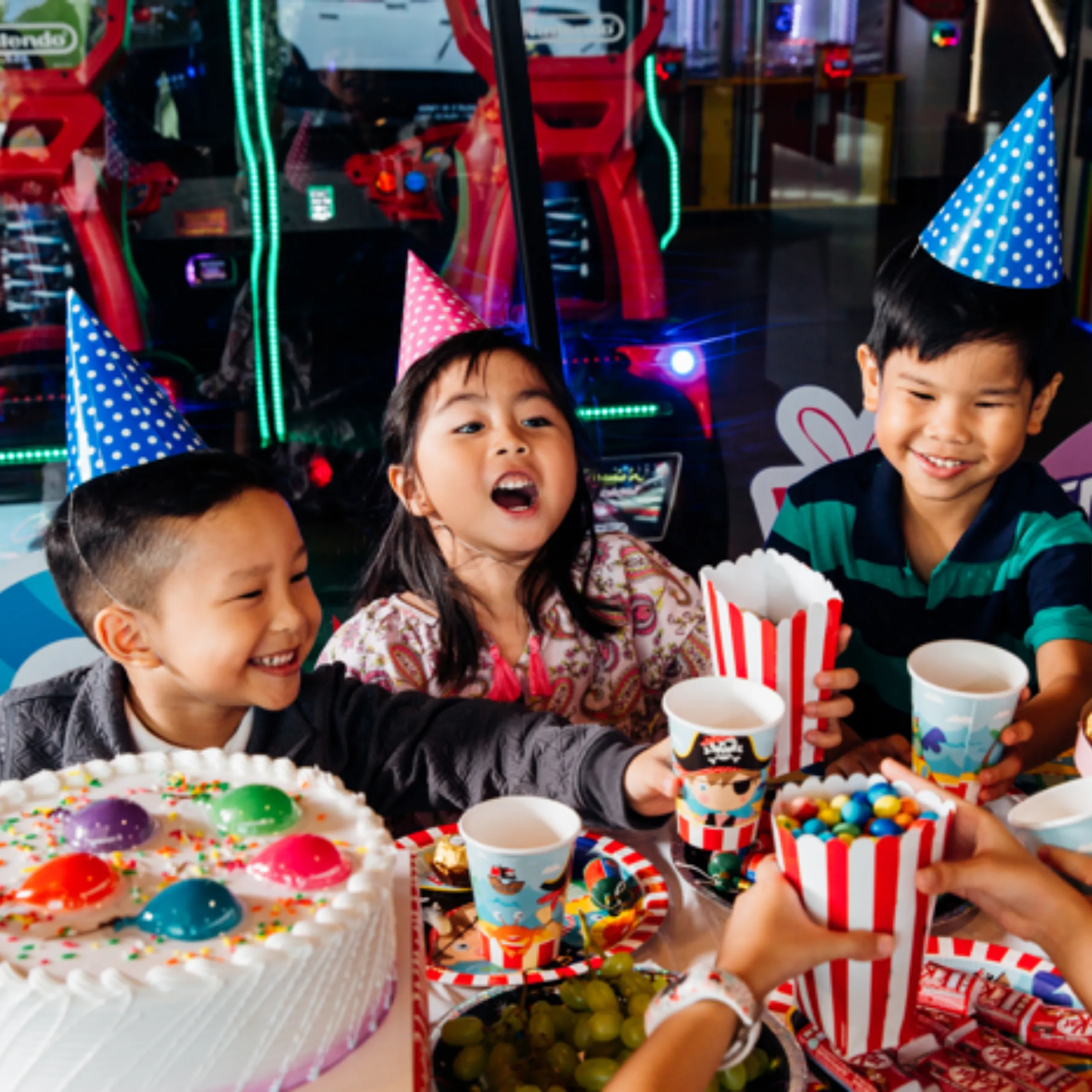 Three smiling children in colorful party hats celebrate at an arcade with a decorated birthday cake, popcorn, drinks, and candy.