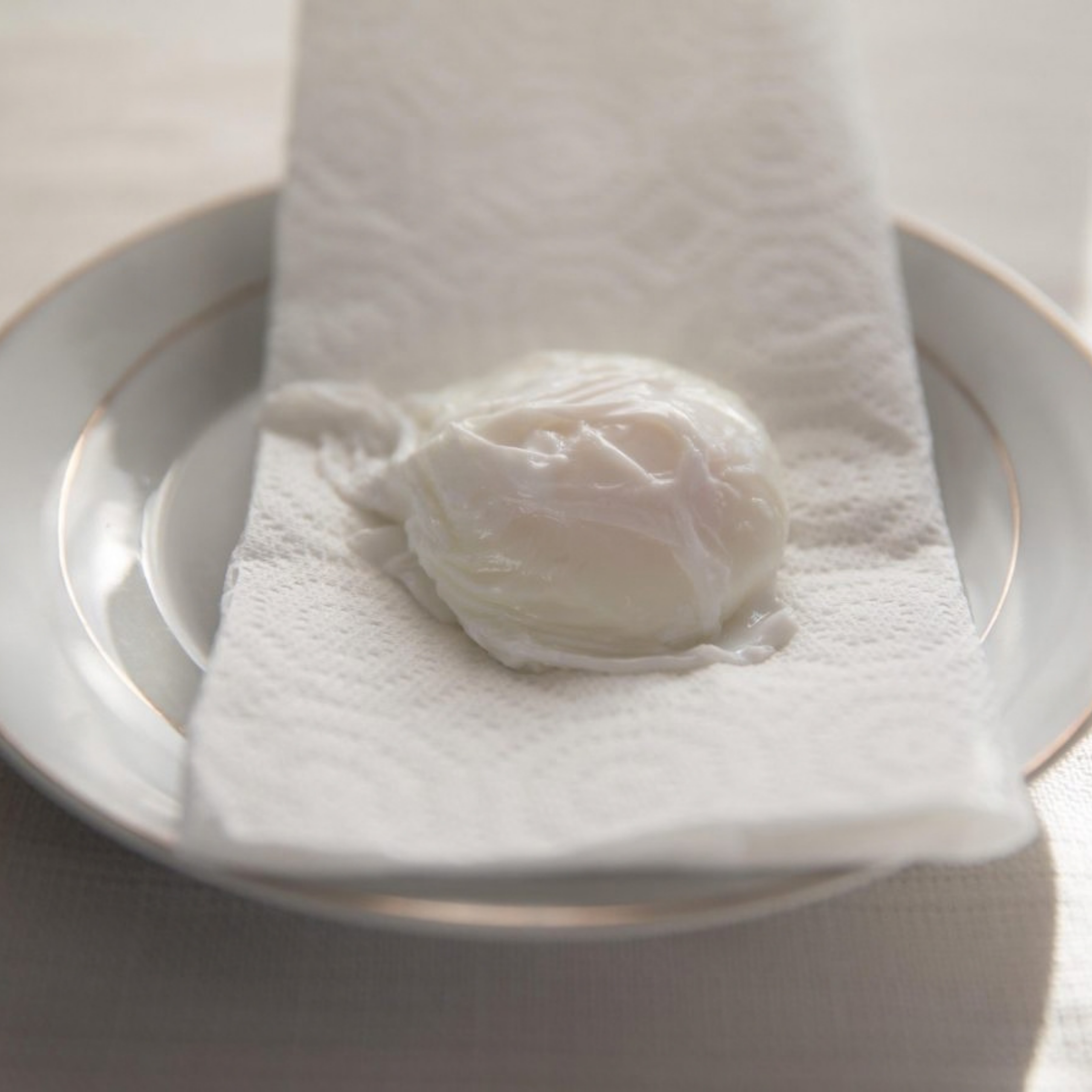 A soft-poached egg rests on a textured white paper towel atop a clear glass plate, lit by gentle natural light for a quiet, minimal kitchen scene.
