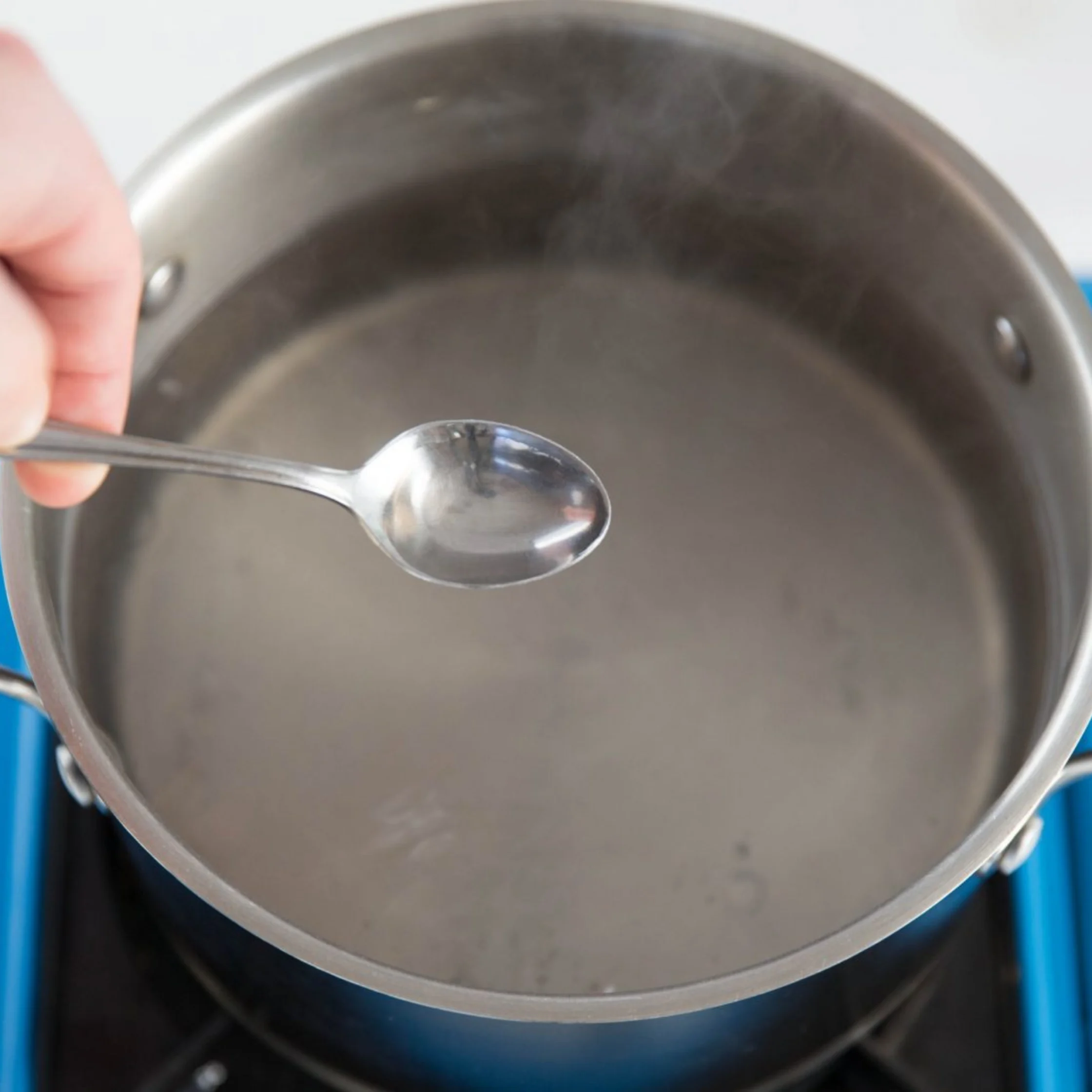 A hand holds a metal spoon over a large stainless-steel pot of clear water on a blue stovetop, suggesting a calm moment of cooking or checking the water.