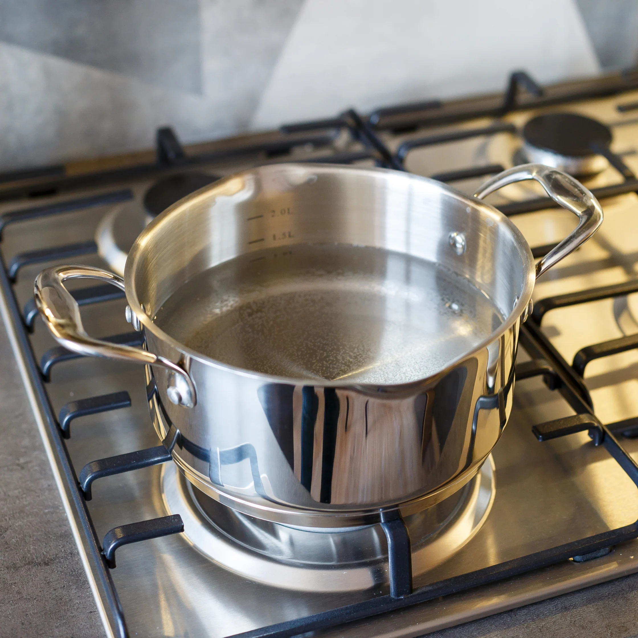 A shiny stainless-steel pot filled with water rests on a gas stovetop, reflecting a clean kitchen with a bold gray-and-white geometric backsplash.