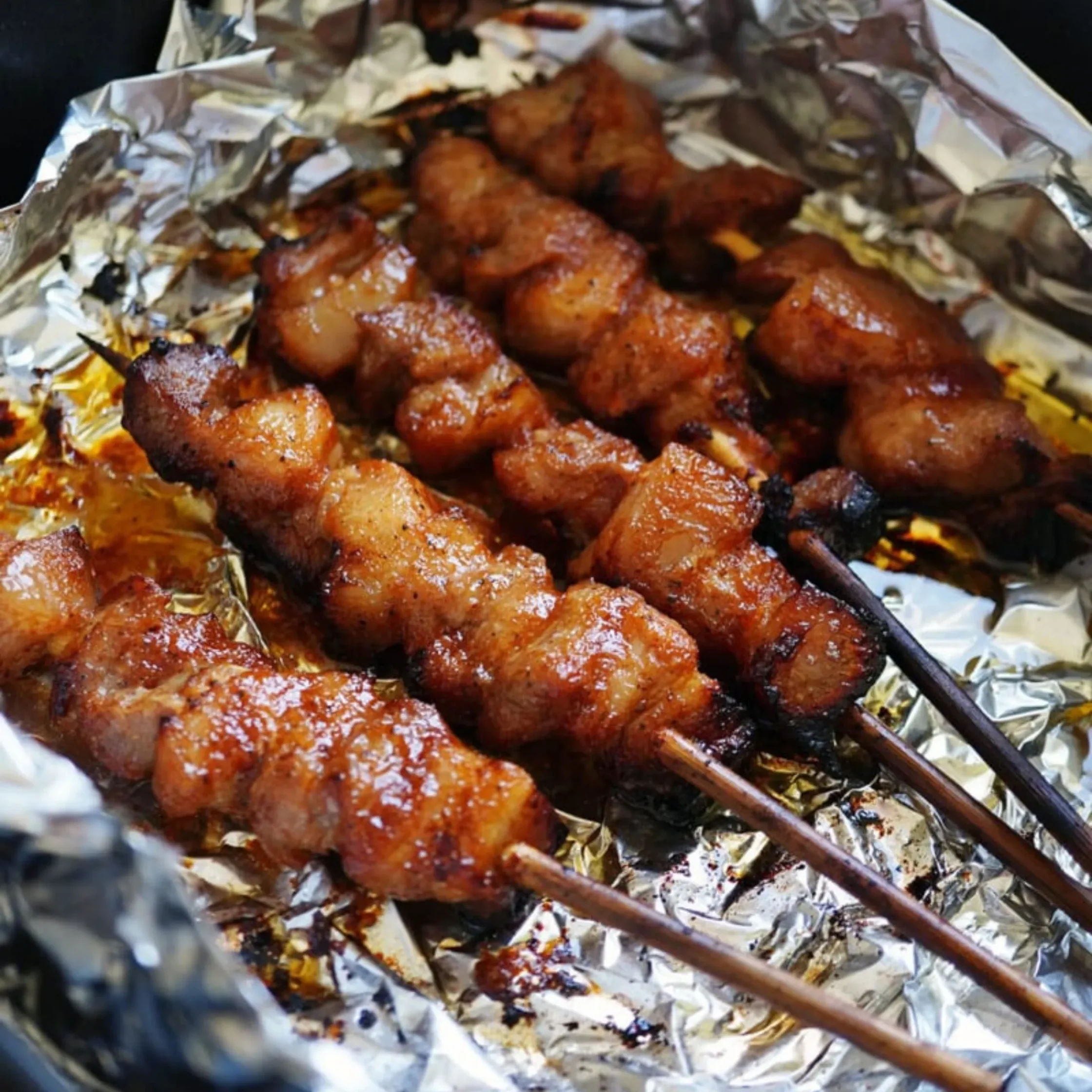 Close-up of golden-brown grilled meat skewers arranged on foil in a black tray, glistening with sauce and conveying a savory, appetizing warmth.