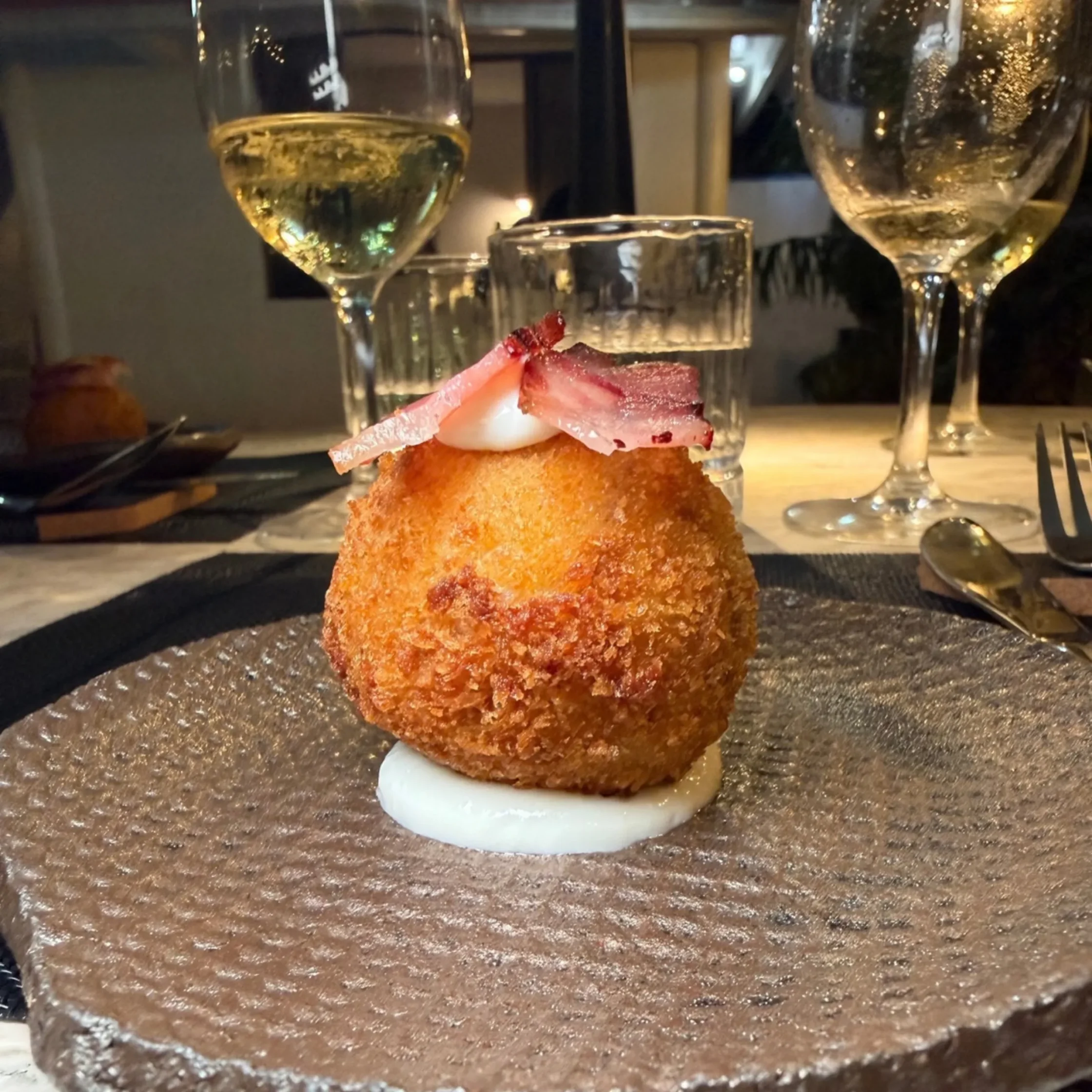 Golden-brown breaded croquette topped with white cream and pink cured ham, served on a textured plate; wine glasses add an elegant restaurant mood.