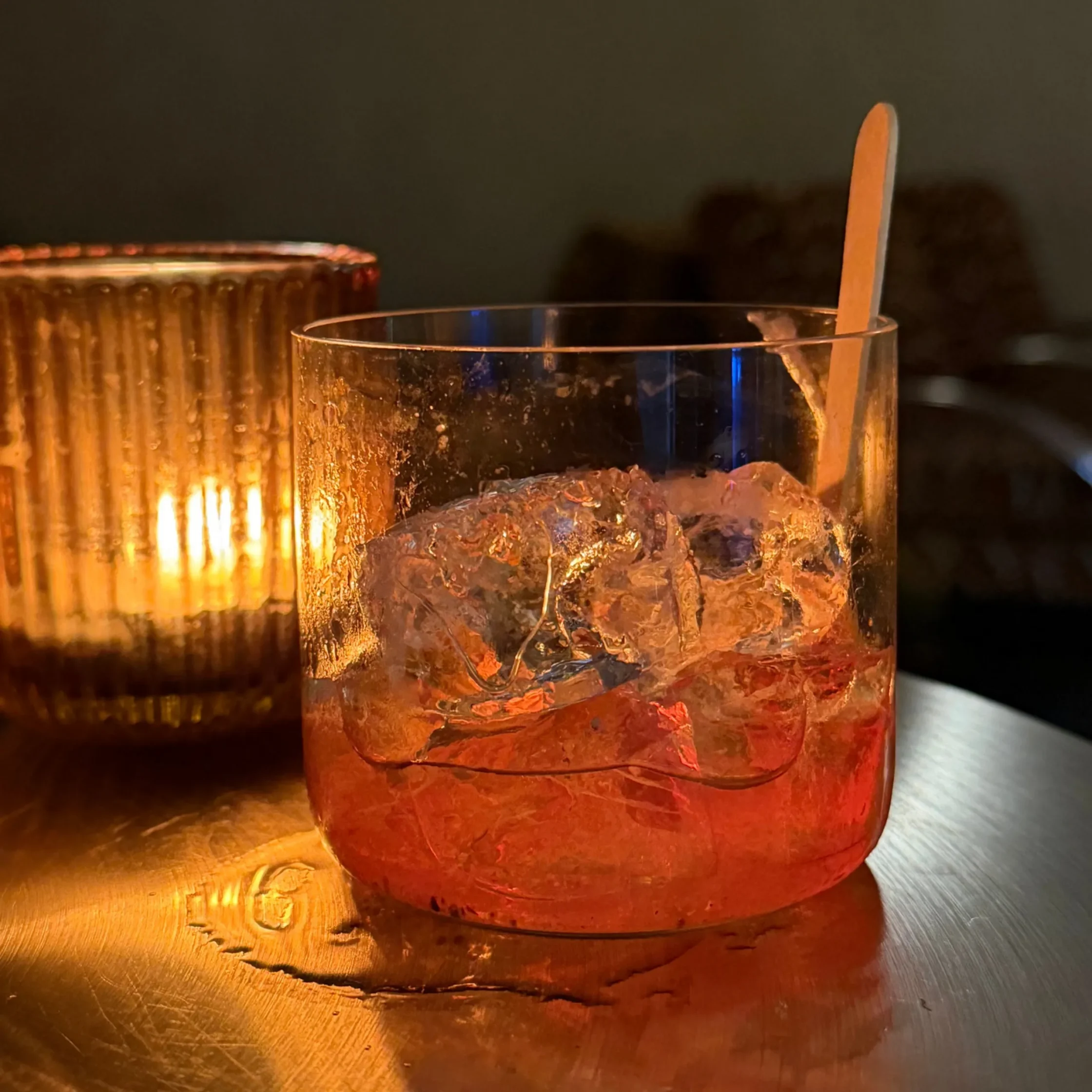 Close-up of a glass with ice and a wooden spoon inside, resting on a warm metallic surface with candlelight in the background.