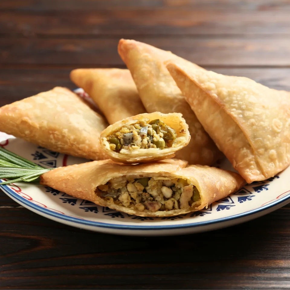 Close-up of golden fried pastry turnovers with a visible chopped vegetable filling on a patterned plate, set against a dark wooden table.