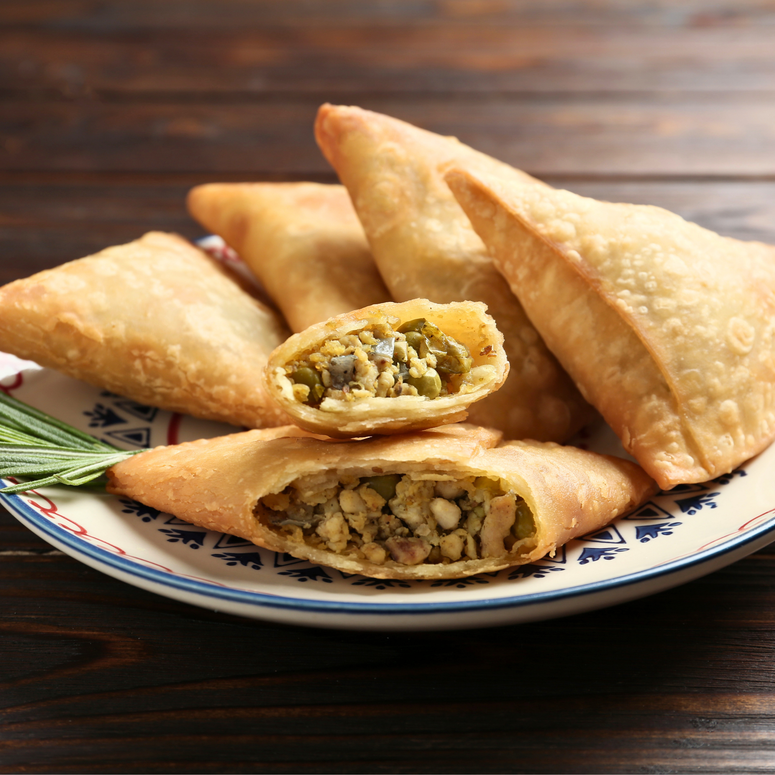 Close-up of golden fried pastry turnovers with a visible chopped vegetable filling on a patterned plate, set against a dark wooden table.
