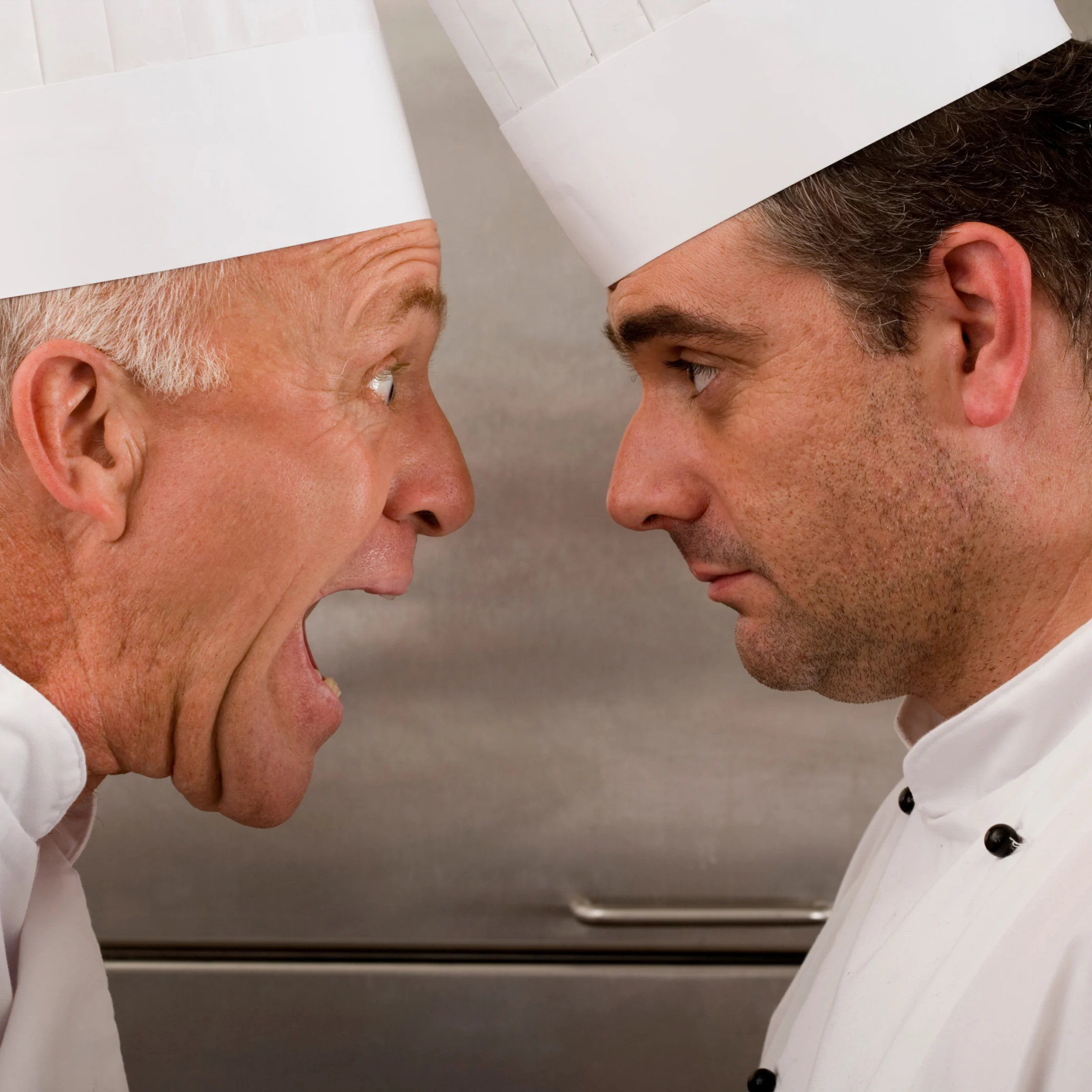 Two chefs in white uniforms and tall hats confront each other face-to-face in a tense kitchen standoff; one shouts, the other looks serious.