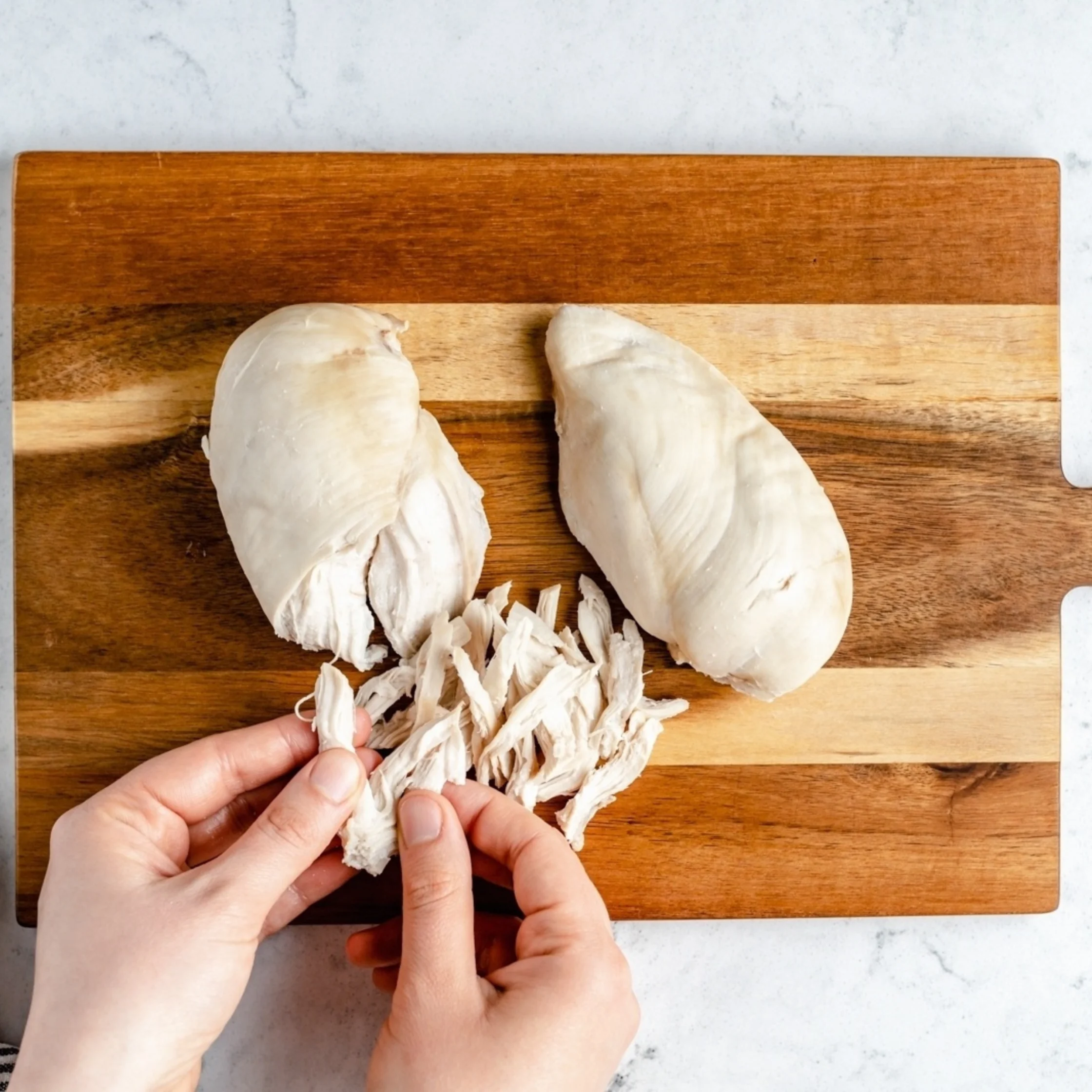 Hands shred cooked chicken breasts on a wooden cutting board, with two poached pieces and pulled strips on a pale marble surface.