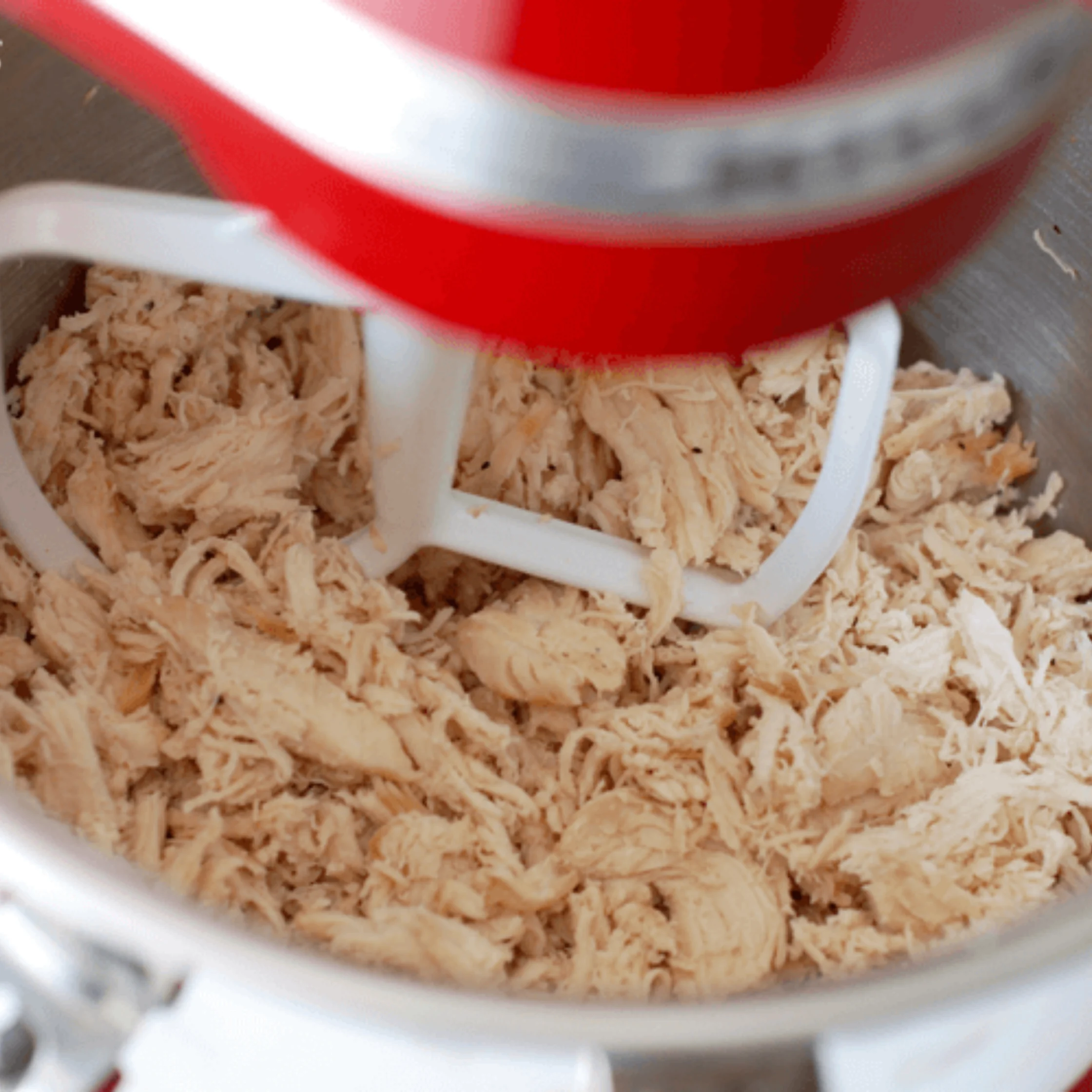 Close-up of a red stand mixer’s white paddle stirring shredded cooked chicken in a metal bowl, capturing an active food-preparation scene.