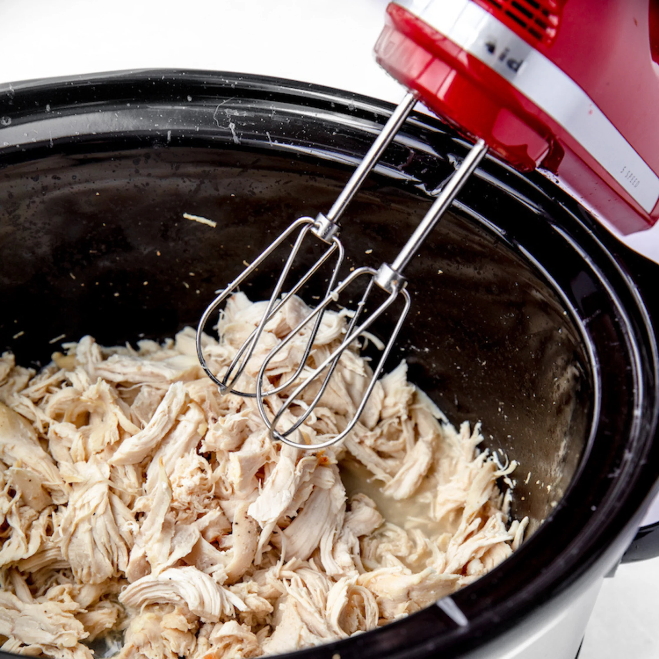 Red-and-silver hand mixer shredding cooked chicken in a black bowl, showing a practical kitchen technique for meal preparation.