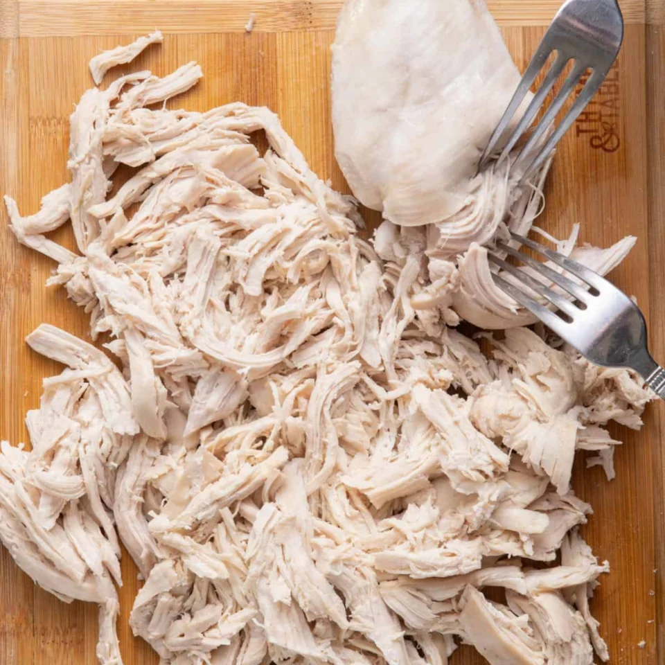 Top-down view of shredded cooked chicken on a wooden cutting board, with a chicken breast and two forks creating a rustic food scene.