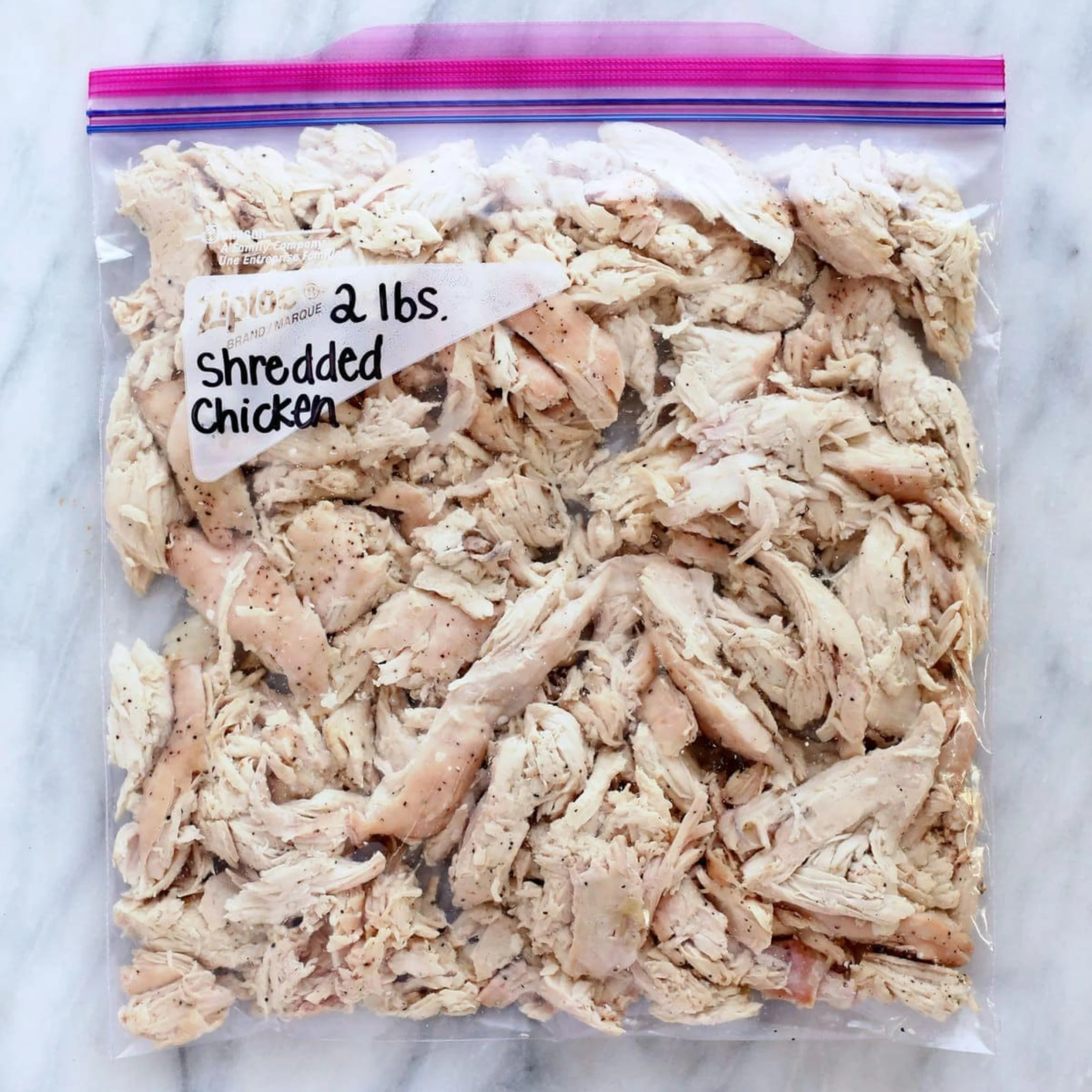 Clear zip-top bag filled with shredded chicken, labeled “2 lbs. Shredded Chicken,” resting on a white marble surface; practical meal-prep image.