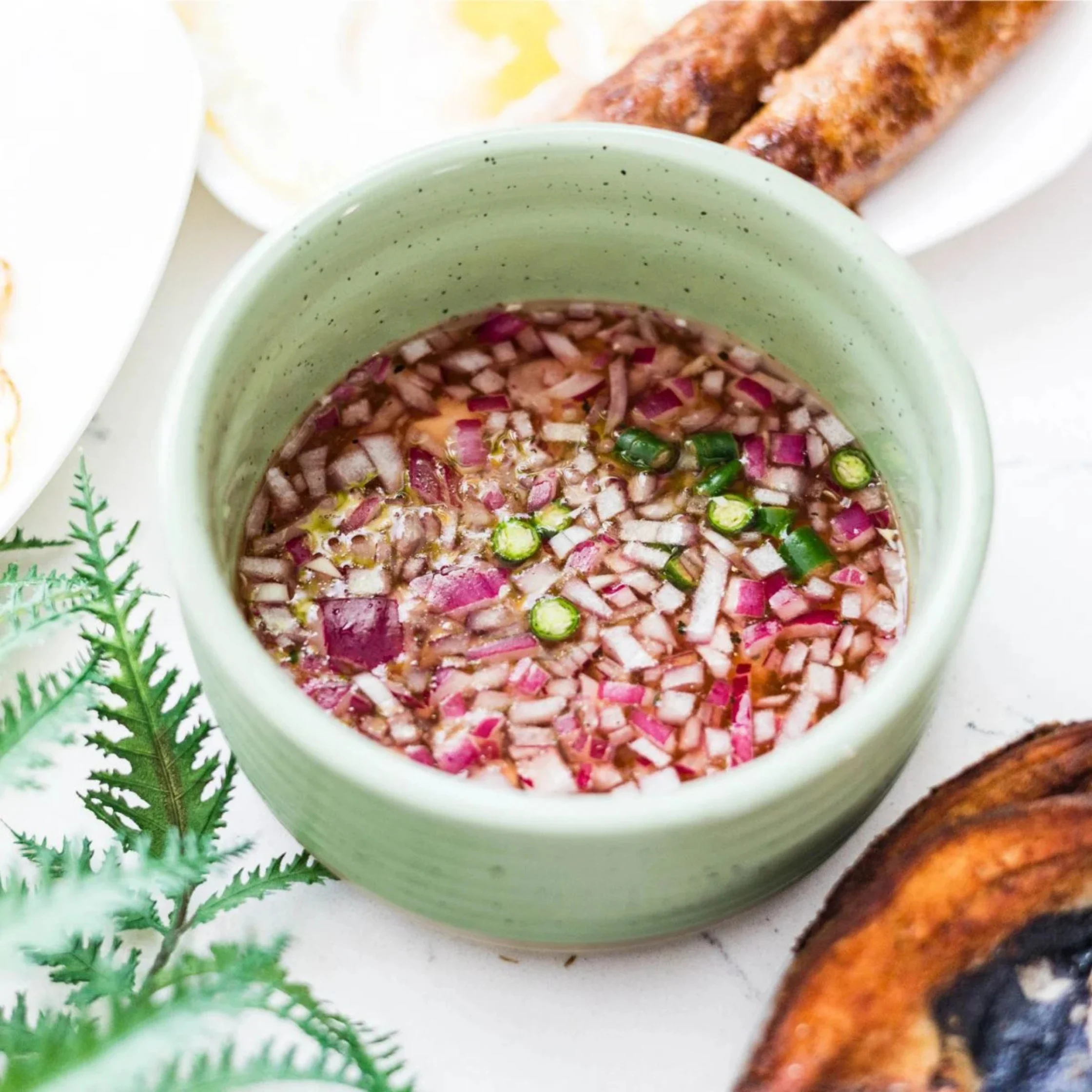 A pale green bowl holds tangy dipping sauce with chopped red onion and green chilies, beside golden-brown grilled fish on a white table. The scene feels fresh and appetizing.