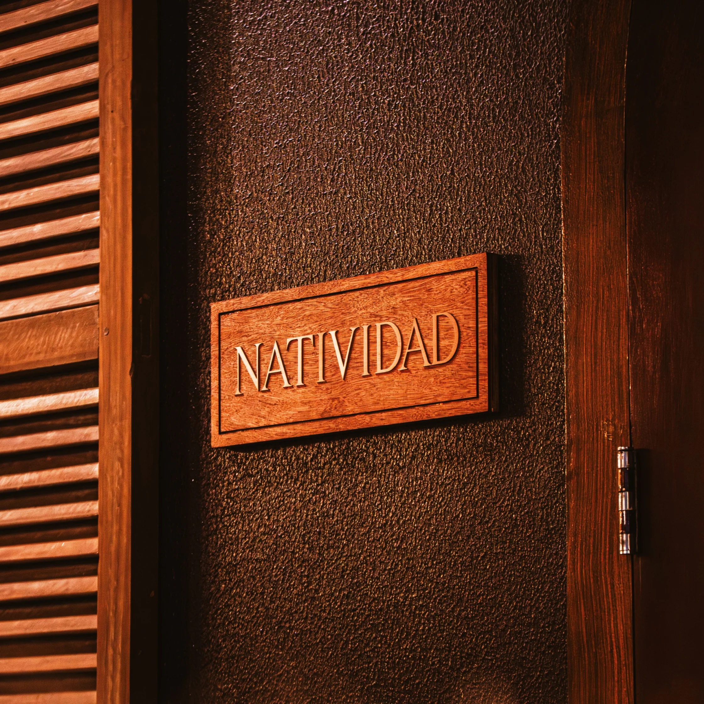 A wooden plaque reading “NATIVIDAD” hangs on a dark, textured door beside wooden slats, lit in warm amber tones that feel welcoming and rustic.