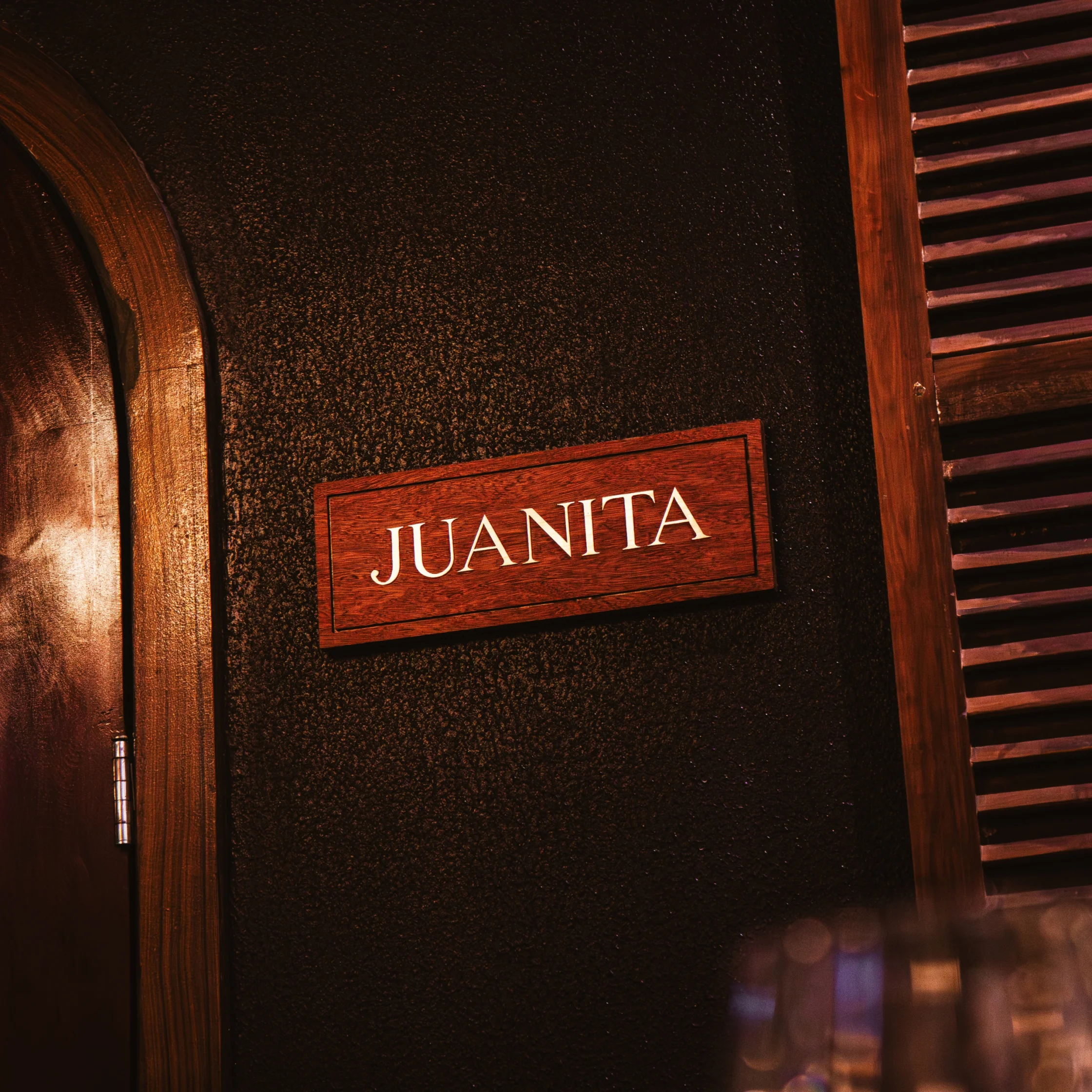 A red wooden plaque reading “JUANITA” hangs on a dark textured wall between an arched door and wooden slats, evoking a warm, intimate mood.