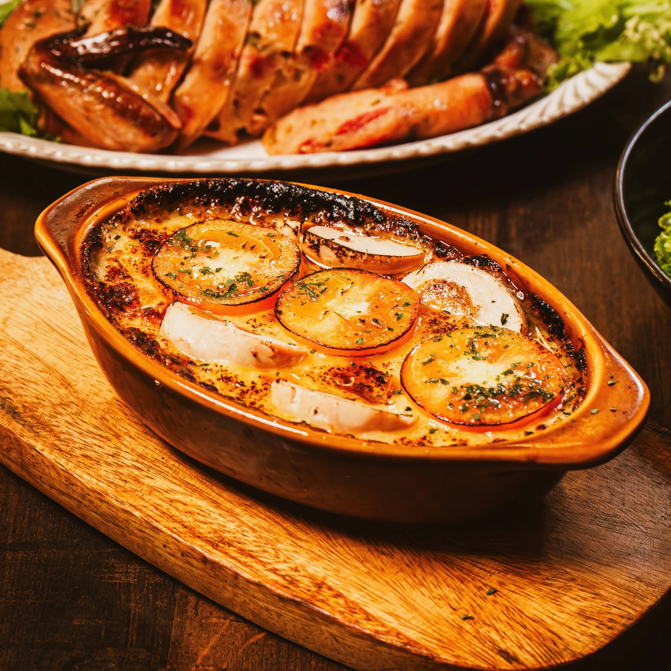 A golden baked tomato and cheese casserole bubbles in an oval dish on a wooden board, with roasted meat and greens behind it; the warm scene feels hearty and inviting.