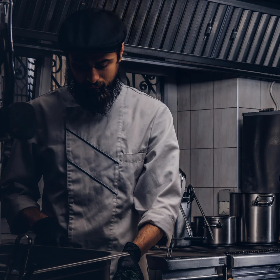 A chef in a black cap and white jacket carefully prepares food in a dim, modern kitchen, creating a focused, moody atmosphere.