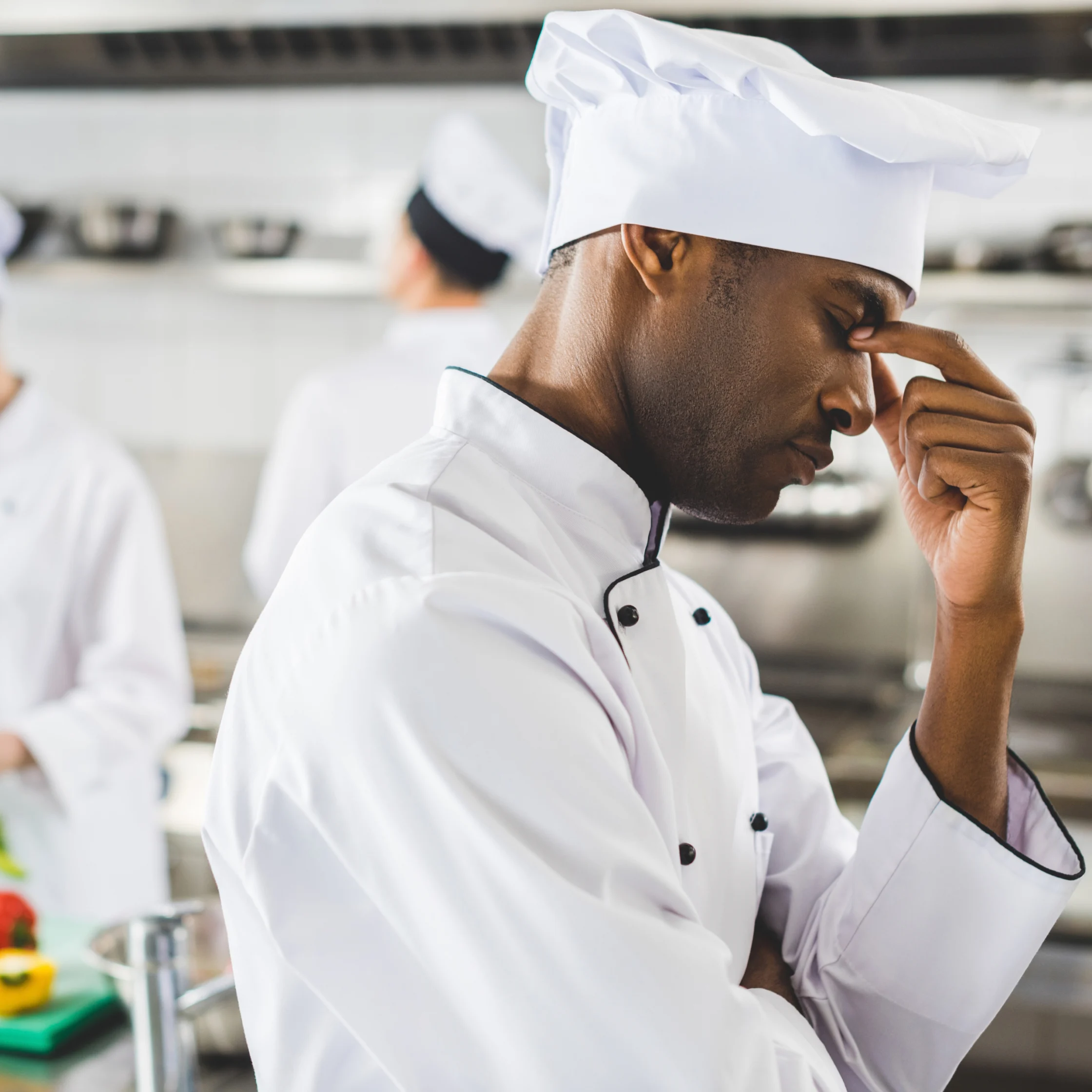 A chef in a white uniform and tall hat pauses thoughtfully in a professional kitchen, touching his forehead as another cook works behind him.