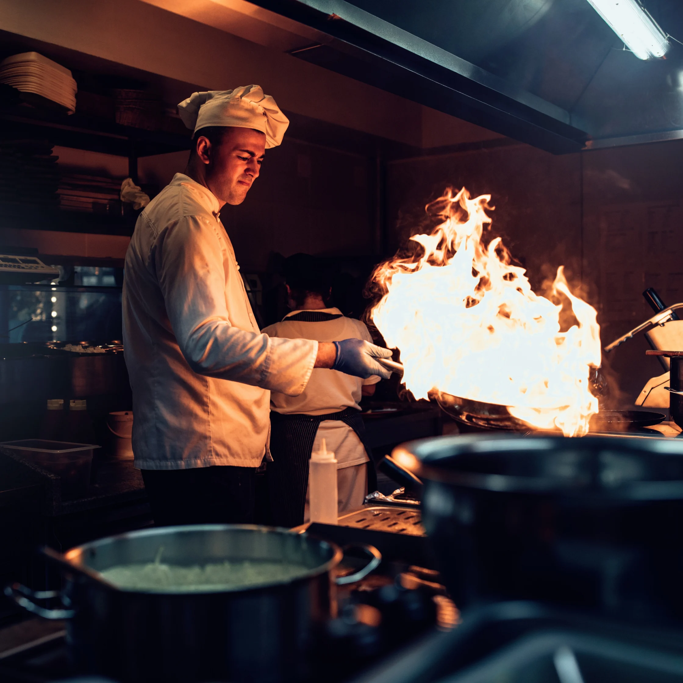 Chef in white uniform cooks at a stove as a dramatic burst of flames rises from a pan in a dim commercial kitchen, creating an intense mood.