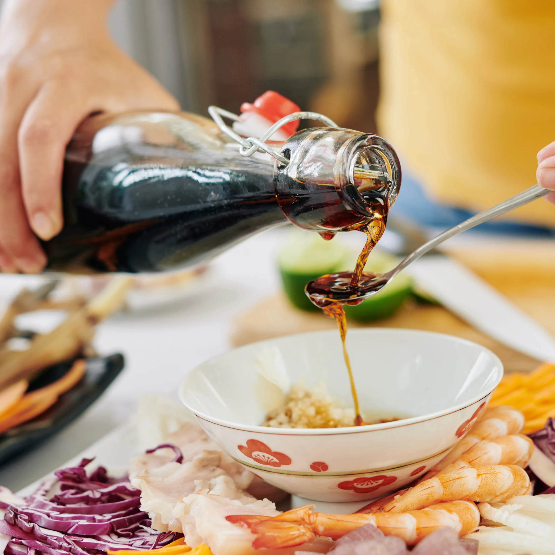 A hand pours dark soy sauce from a glass bottle into a spoon over a bowl of minced garlic, amid shrimp and sliced vegetables; warm, appetizing kitchen scene.