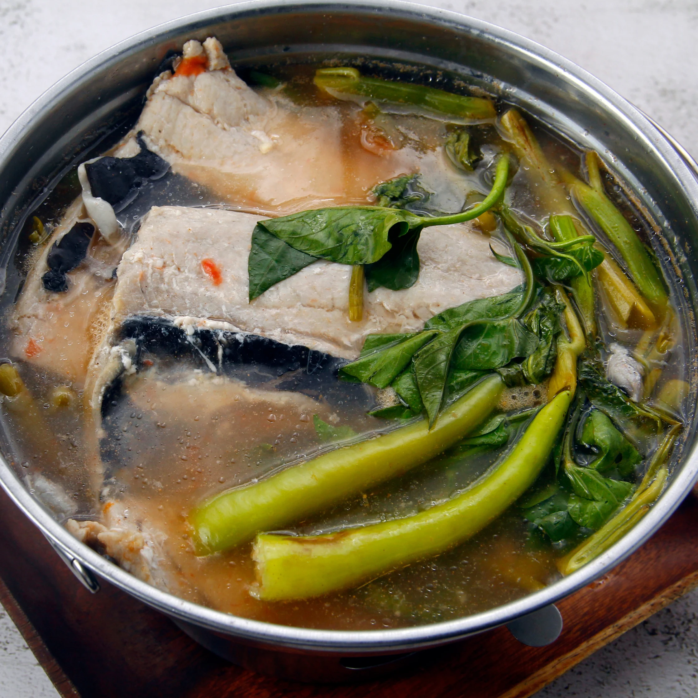 A stainless-steel pot holds clear brown soup with pale fish pieces, leafy greens, and long green stems, evoking a hearty, comforting meal.