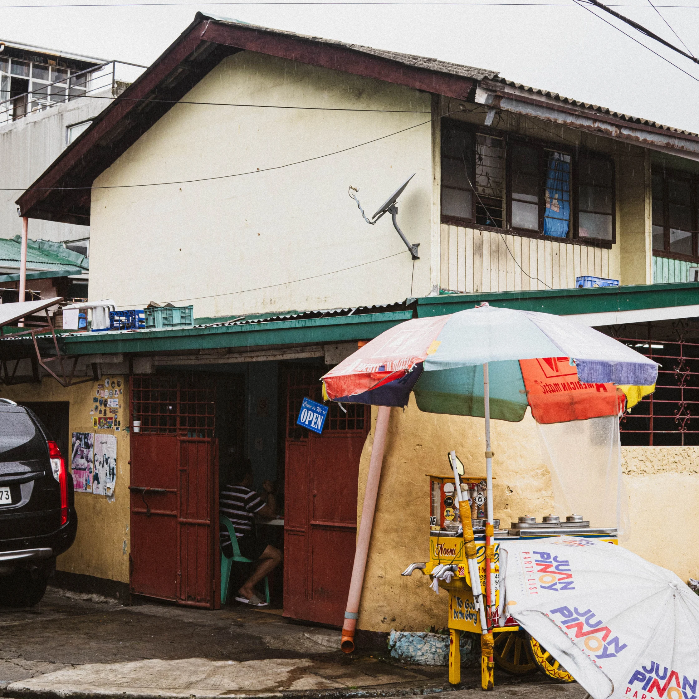 Exterior view of a small shopfront with yellow walls, brown entrance, and a satellite dish on the wall amid power lines.