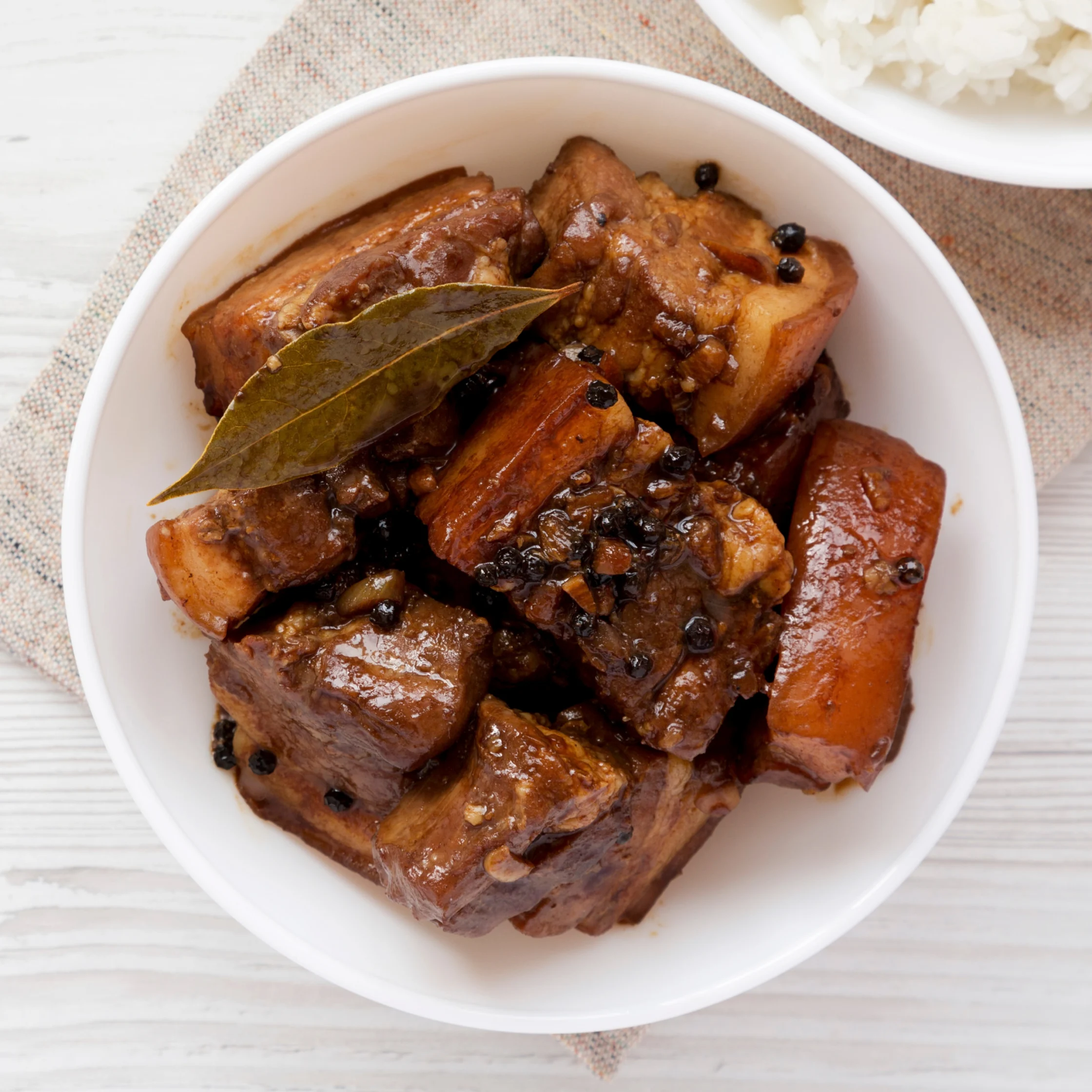 A white bowl of glossy braised pork belly in dark sauce, topped with bay leaves and peppercorns, sits beside white rice for a hearty, comforting meal.