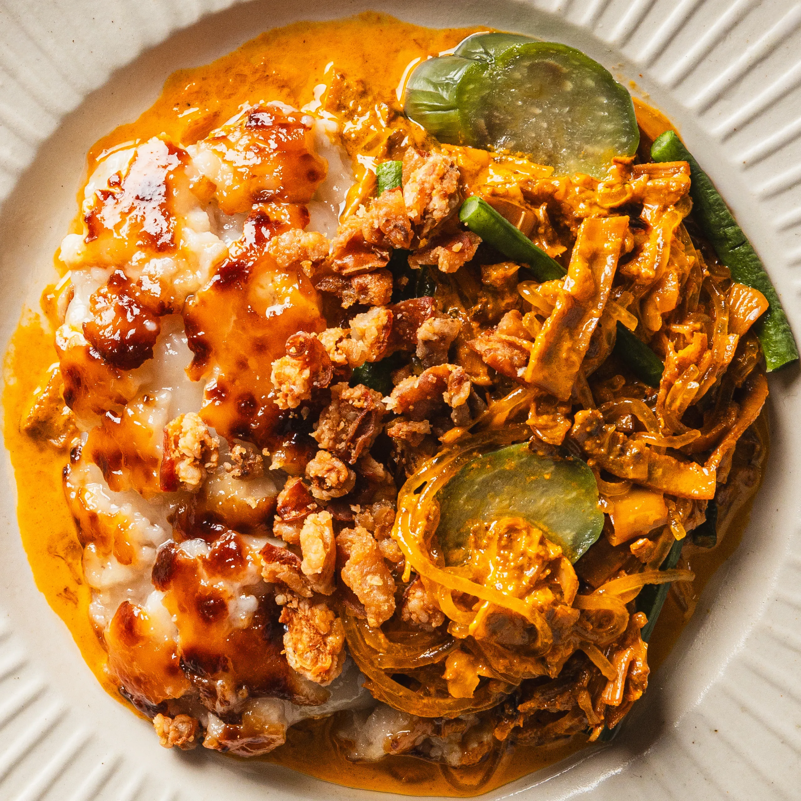 Overhead view of a white bowl filled with spicy orange-sauced noodles, minced meat, sliced pickles, greens, and melted cheese; hearty, vibrant dish.
