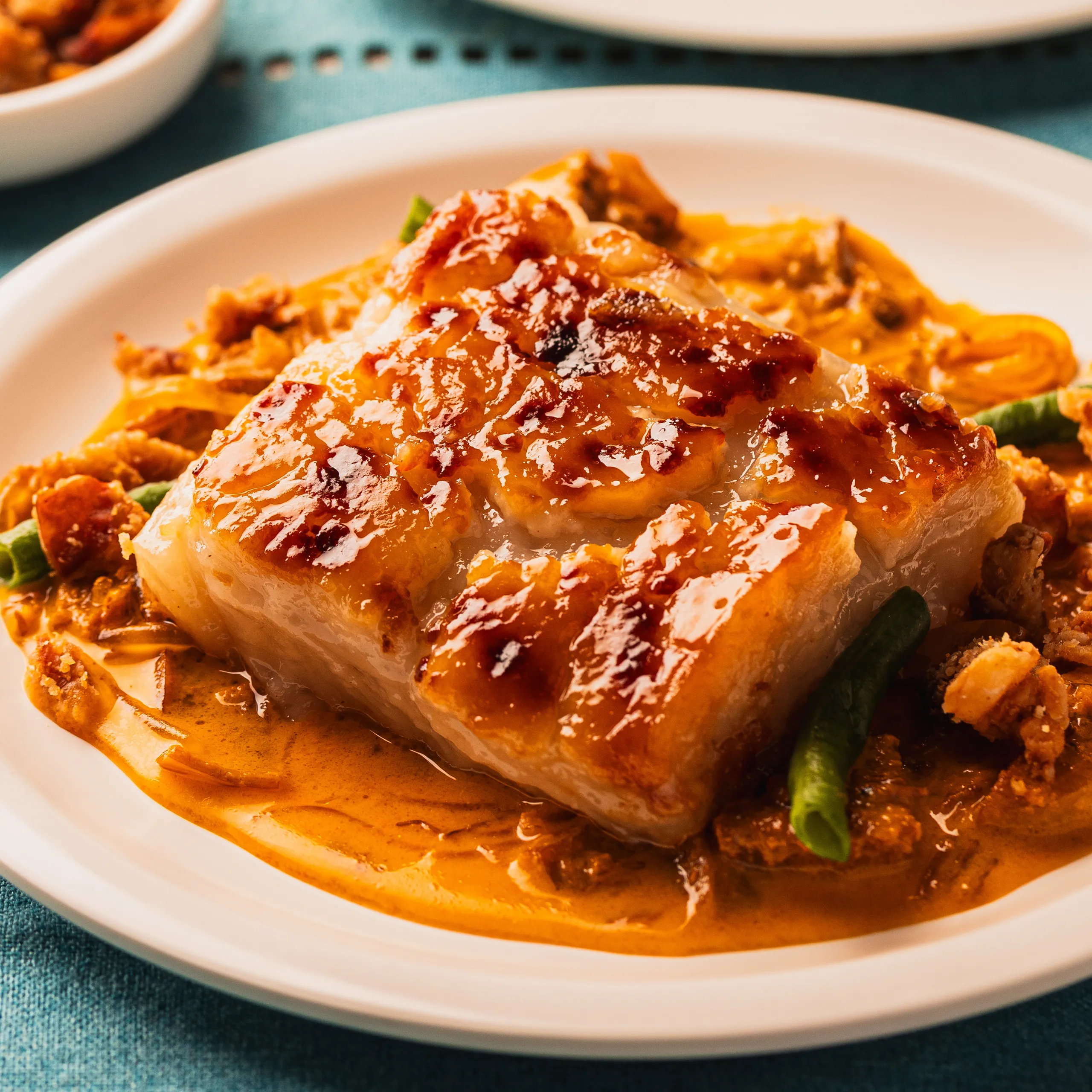 A glossy, golden-brown glazed chicken breast sits in rich orange sauce with vegetables on a white plate, creating a warm, appetizing meal.