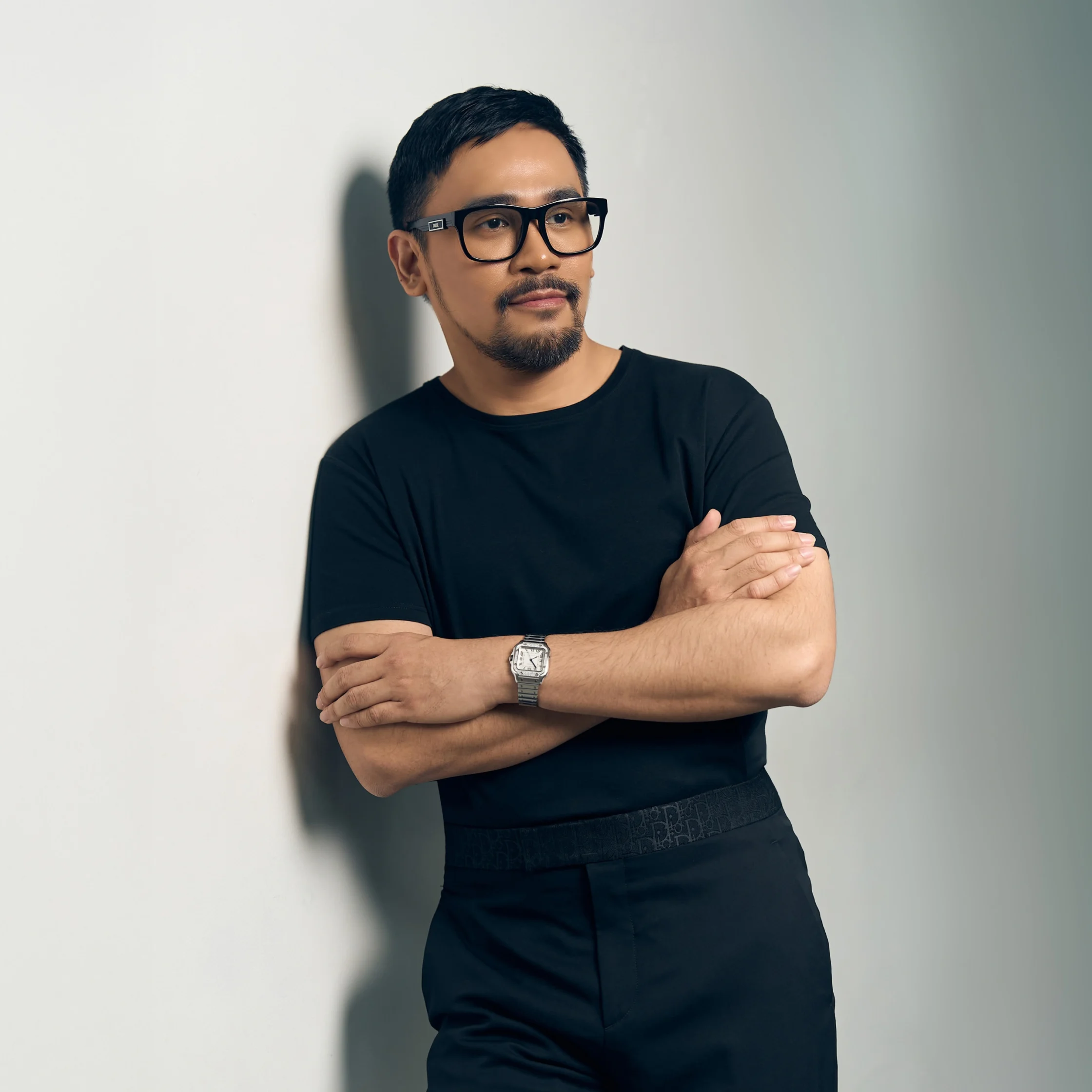 A thoughtful man in black glasses and a dark outfit stands with arms crossed against a light gray wall, conveying a calm, confident mood.