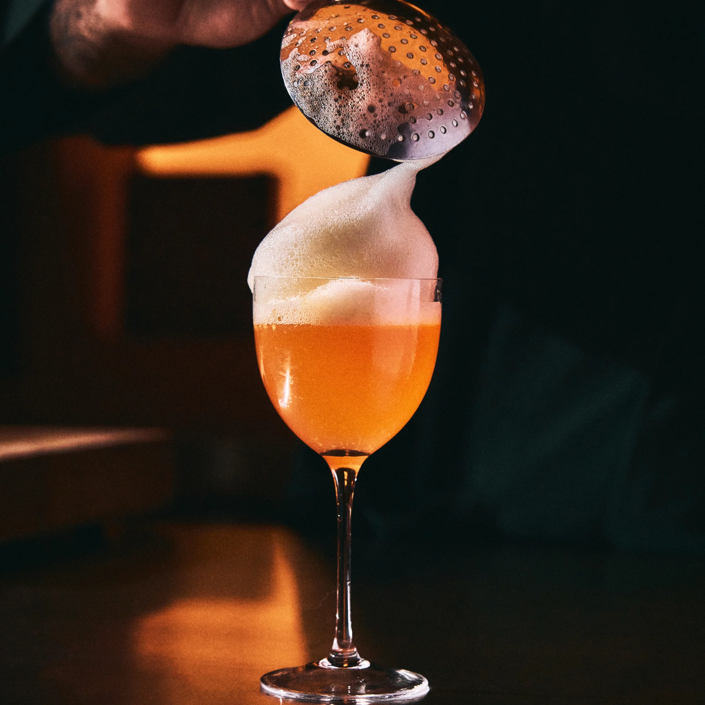 A bartender spoons thick foam onto a stemmed glass of glowing amber cocktail, creating a dramatic pour in a dim, warmly lit bar.