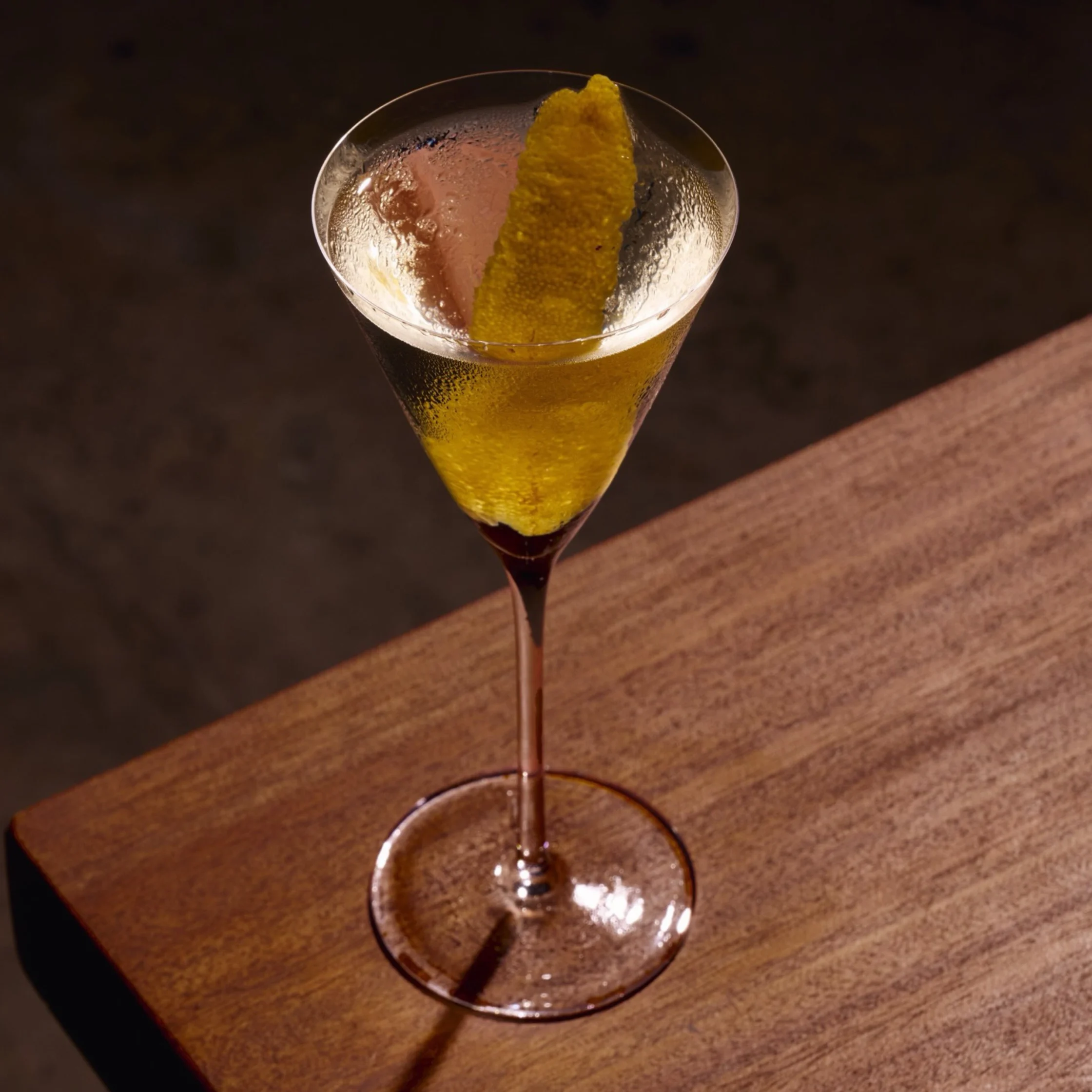 A golden cocktail with a long yellow citrus peel sits in a stemmed martini glass on a wooden bar, lit warmly against a dark background.
