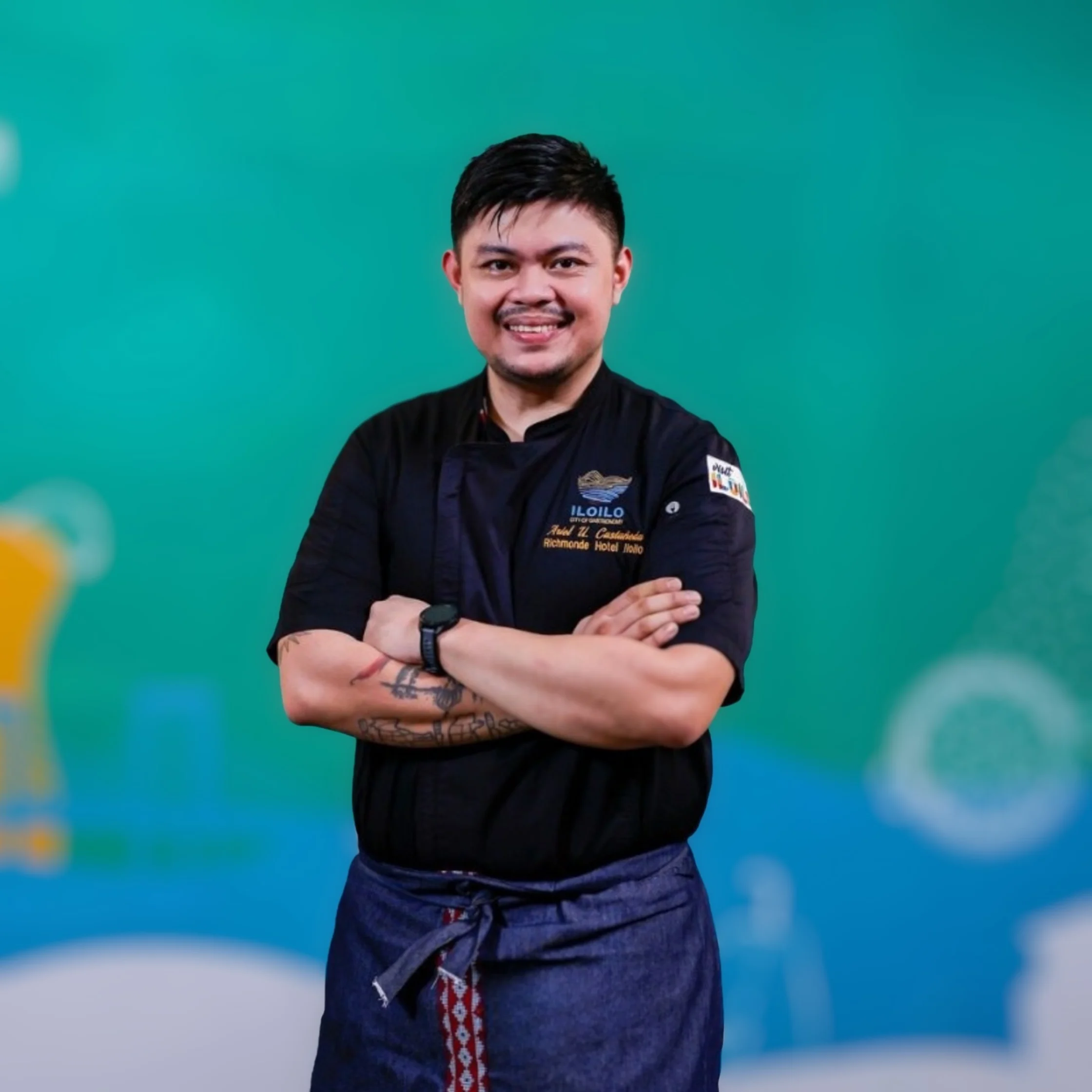 Smiling male chef in a black uniform and blue apron stands with arms crossed against a turquoise and blue backdrop, conveying confidence and warmth.