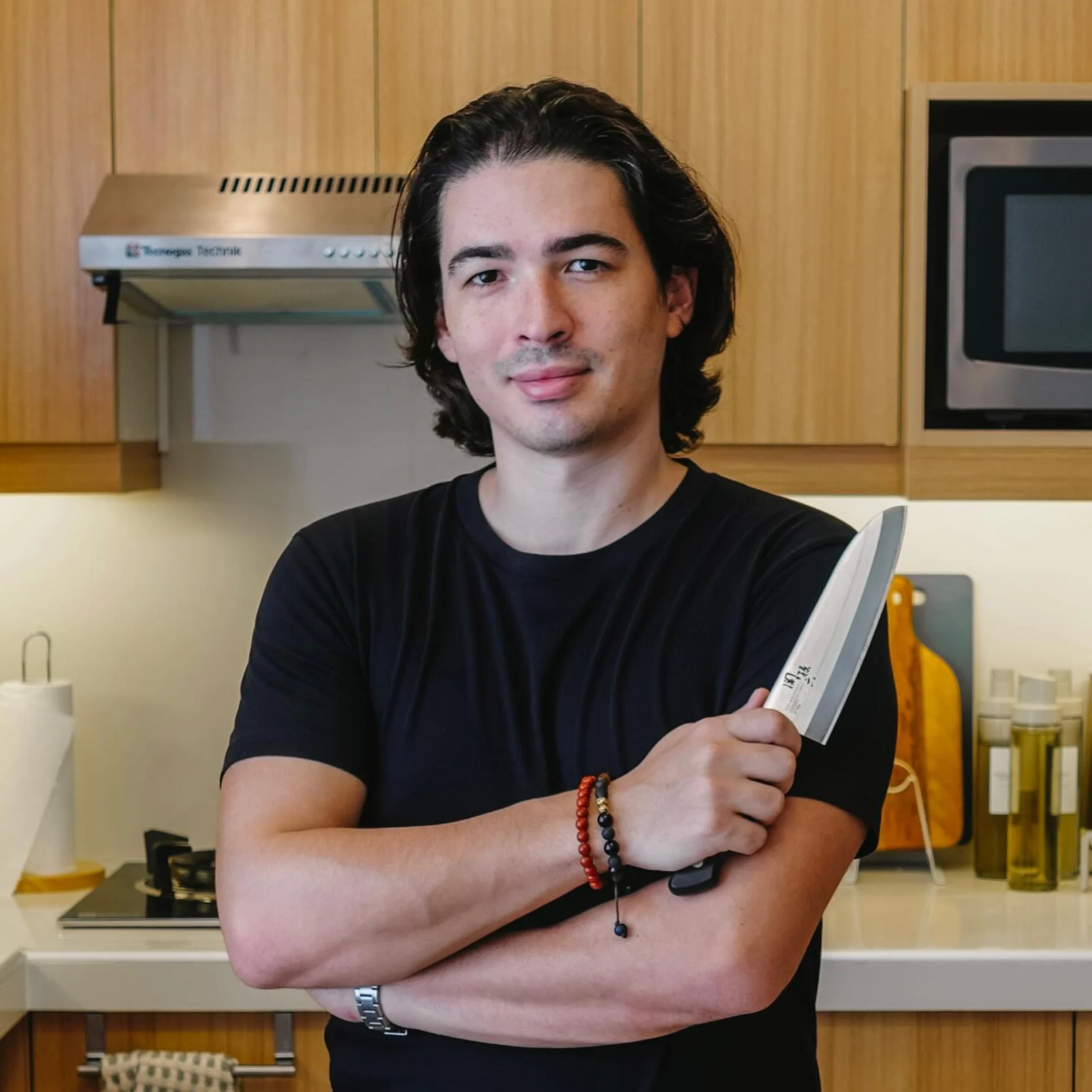 A calm-looking man in a black shirt stands in a home kitchen, holding a chef’s knife across his crossed arms, with wooden cabinets behind him.