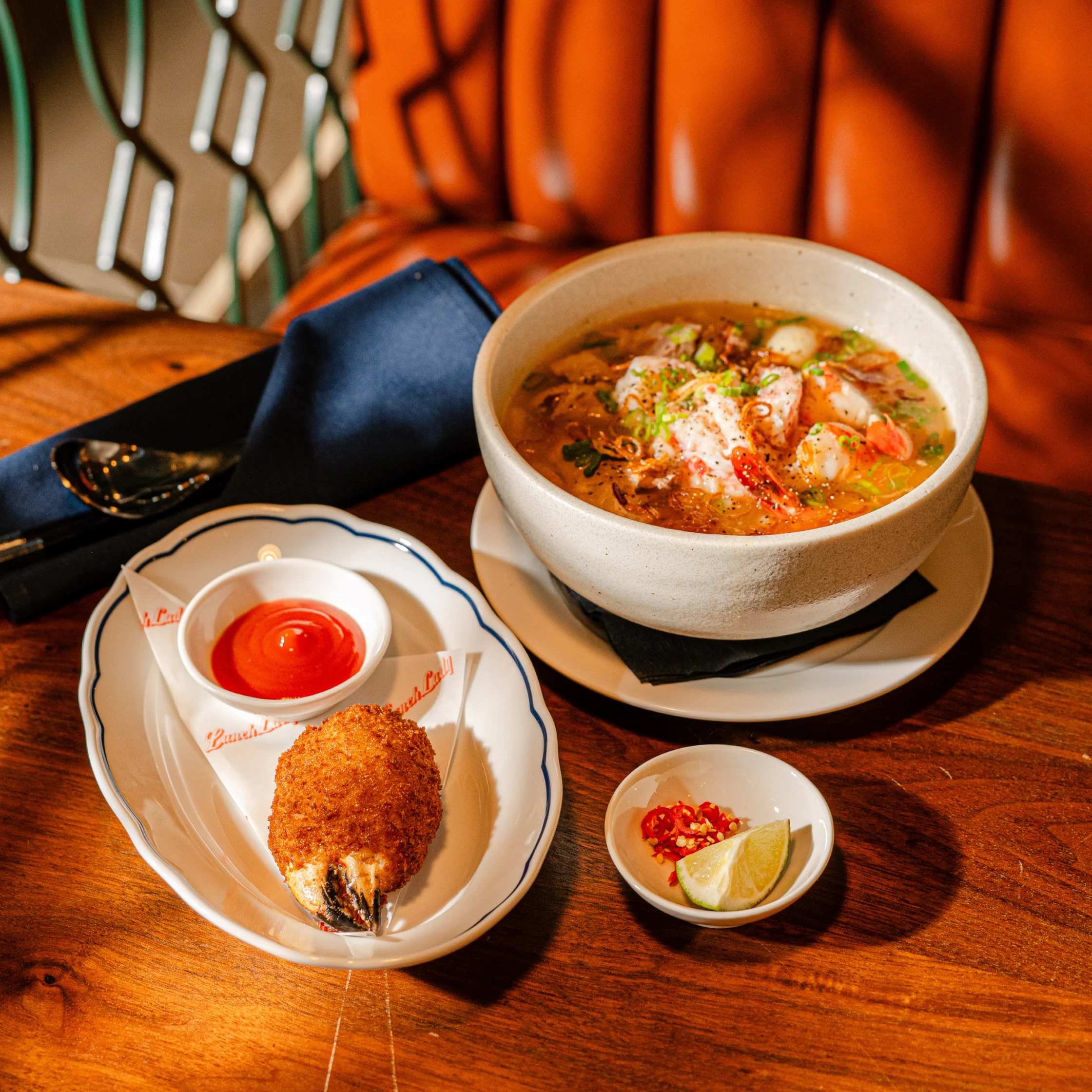 A steaming bowl of spicy seafood soup with shrimp and herbs sits beside a breaded fish cutlet, red dipping sauce, lime, and chili on a wooden table.