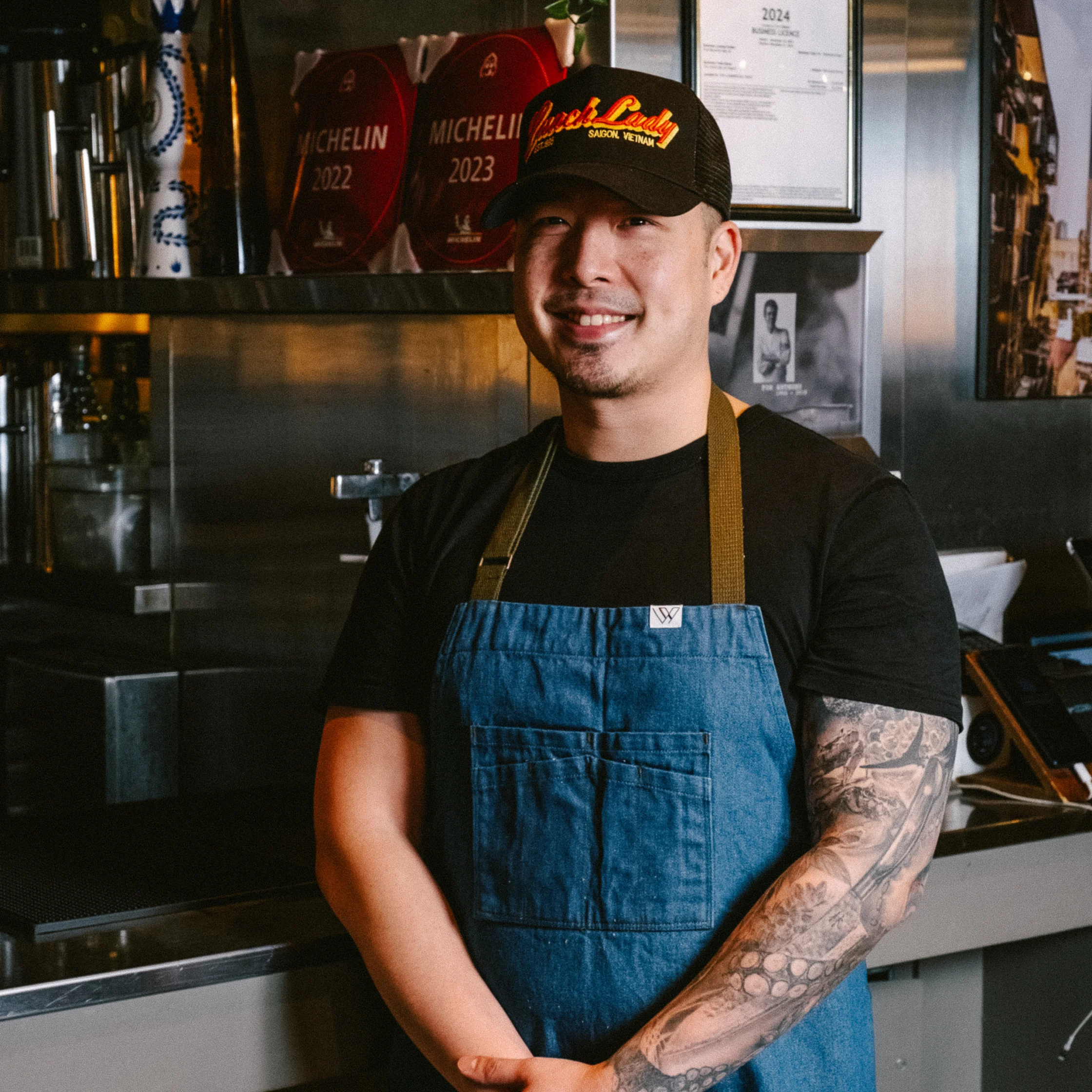 Smiling chef in a black cap and blue apron stands in a restaurant kitchen, with award plaques and framed certificates behind him, conveying pride and warmth.