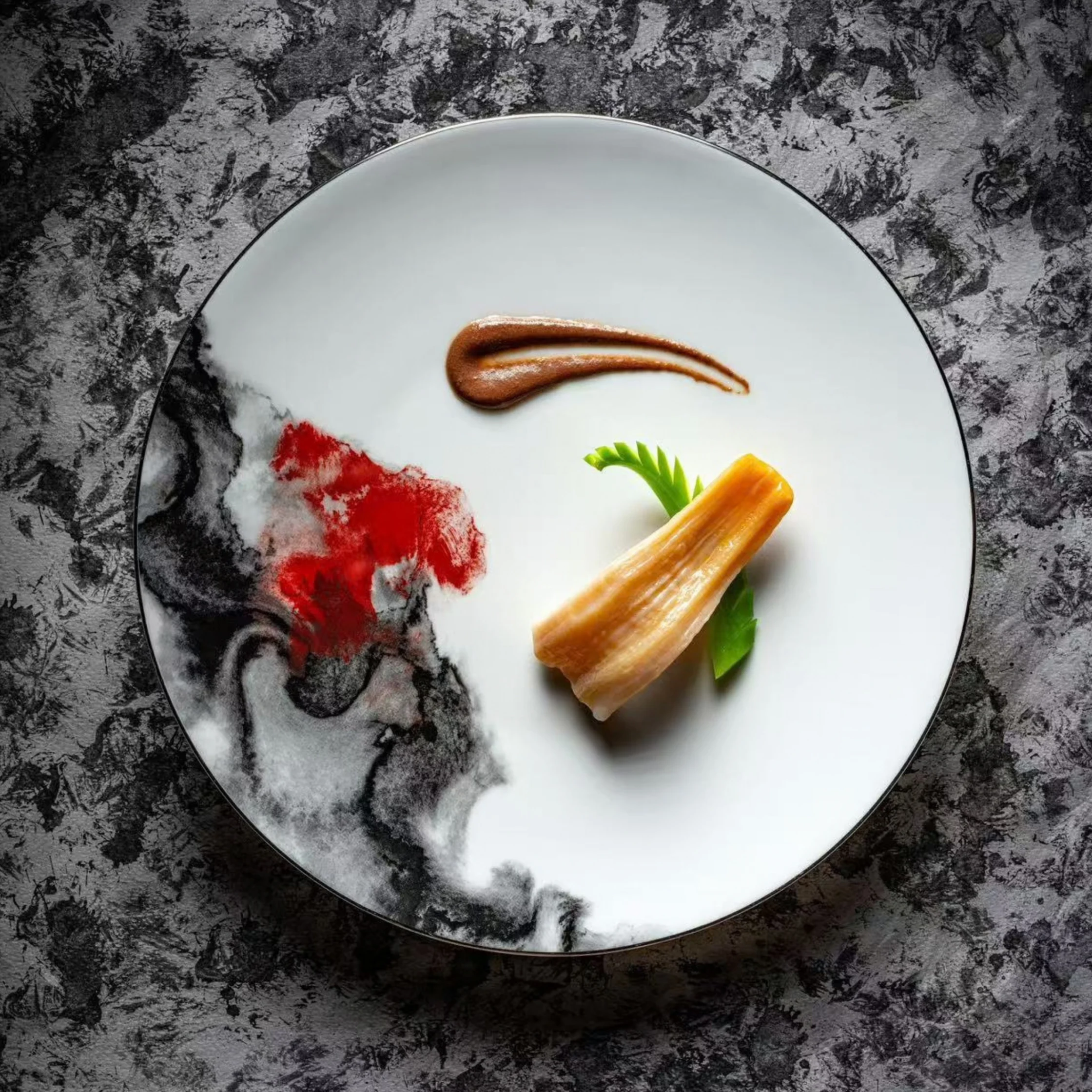 Minimalist gourmet plating: a golden rectangular bite with fresh green leaves and brown sauce sits on a white plate with a red-and-black abstract design, on dark stone.