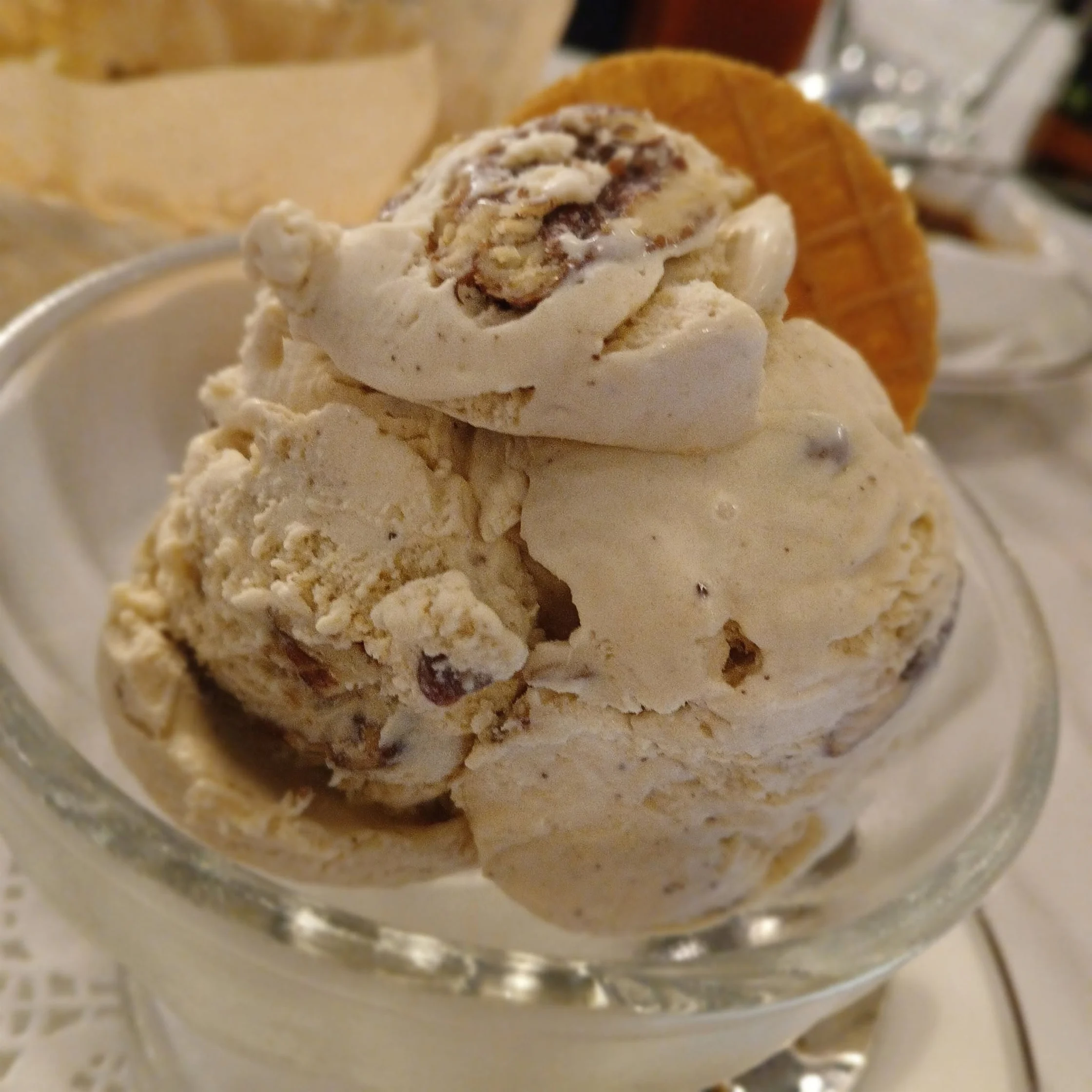 A pink paper cup holds several scoops of creamy vanilla ice cream with golden mango swirls, paired with a purple spoon on a napkin; a cheerful treat.