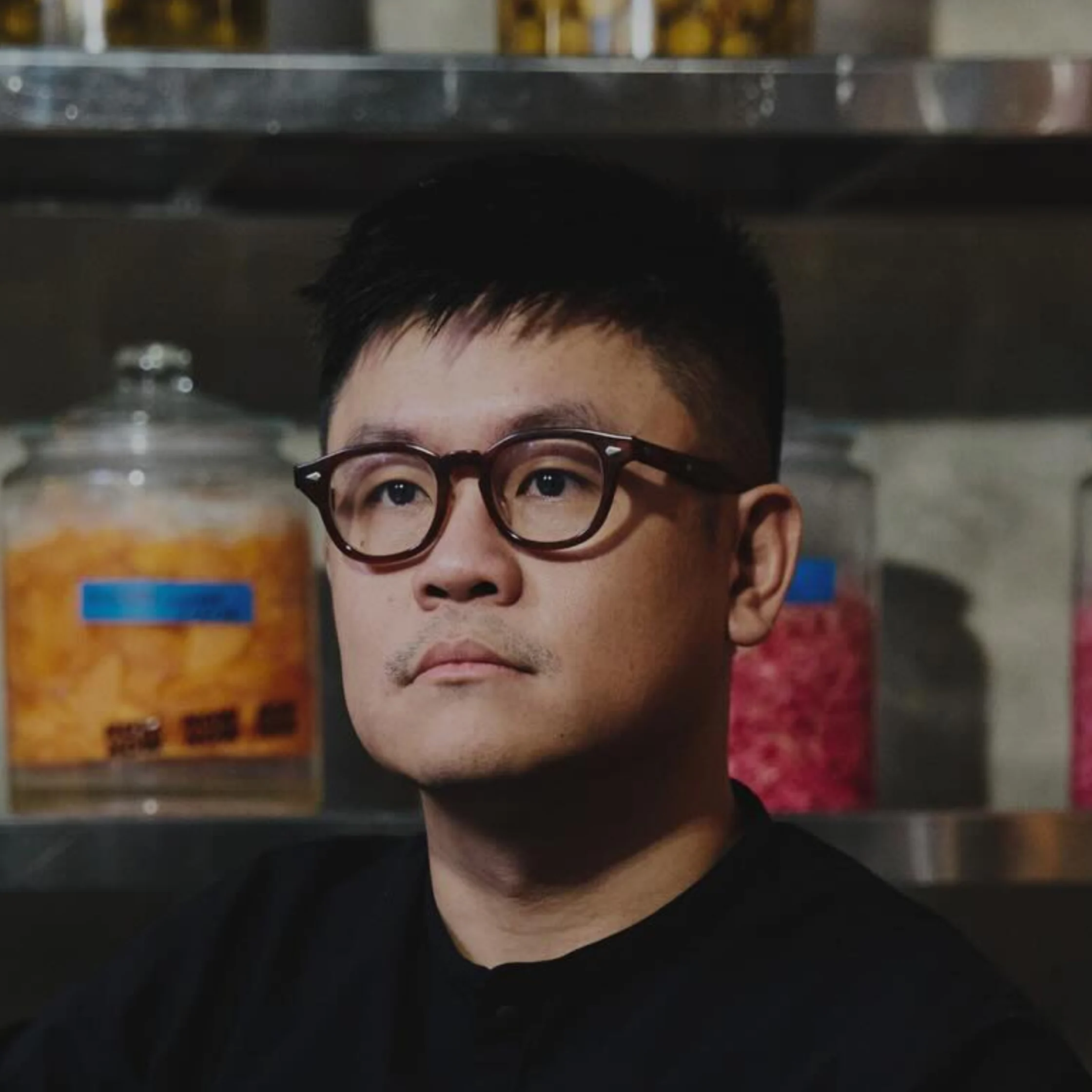 A serious-looking man in dark glasses and a black shirt stands before metal shelves of large jars filled with colorful contents in a dim interior.