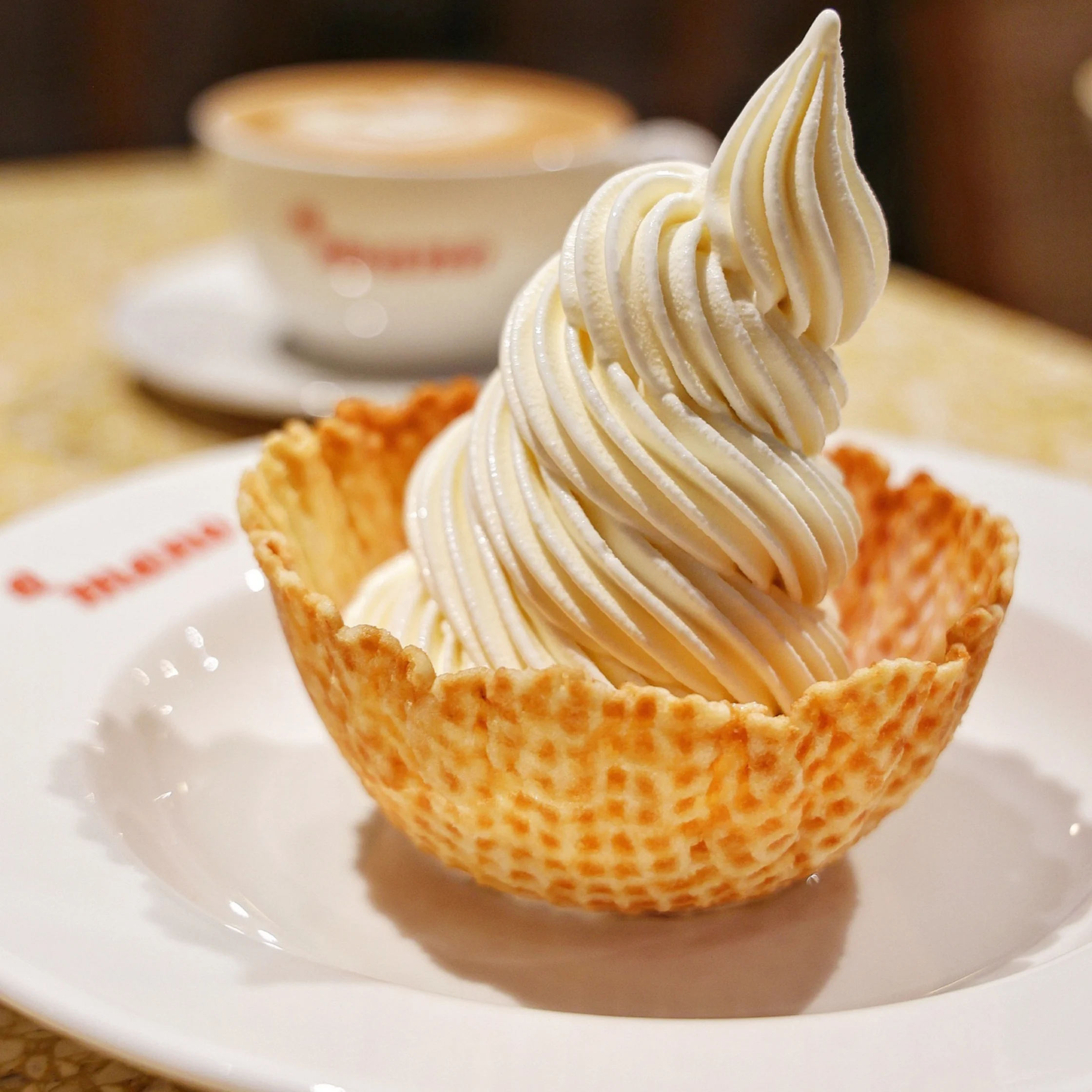 A tall swirl of creamy vanilla soft-serve sits in a crisp waffle bowl on a white plate, with a blurred coffee cup behind it, creating a warm café feel.