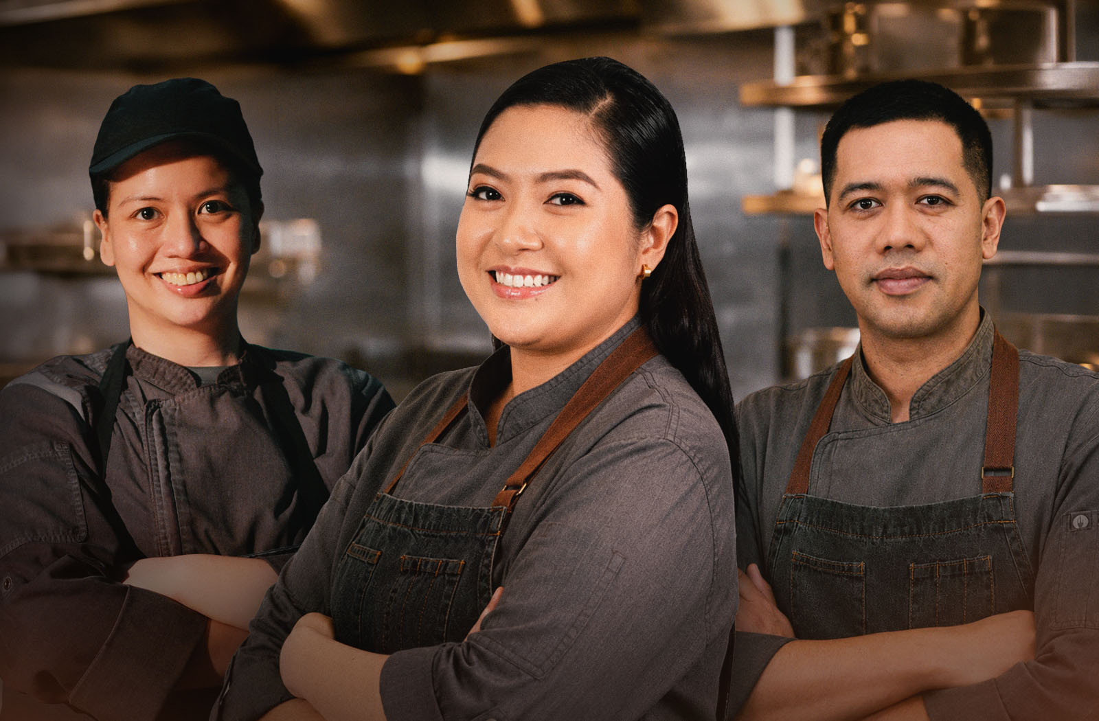 Three restaurant kitchen staff in dark aprons stand with arms crossed, smiling confidently against a stainless-steel kitchen backdrop, conveying teamwork and pride.