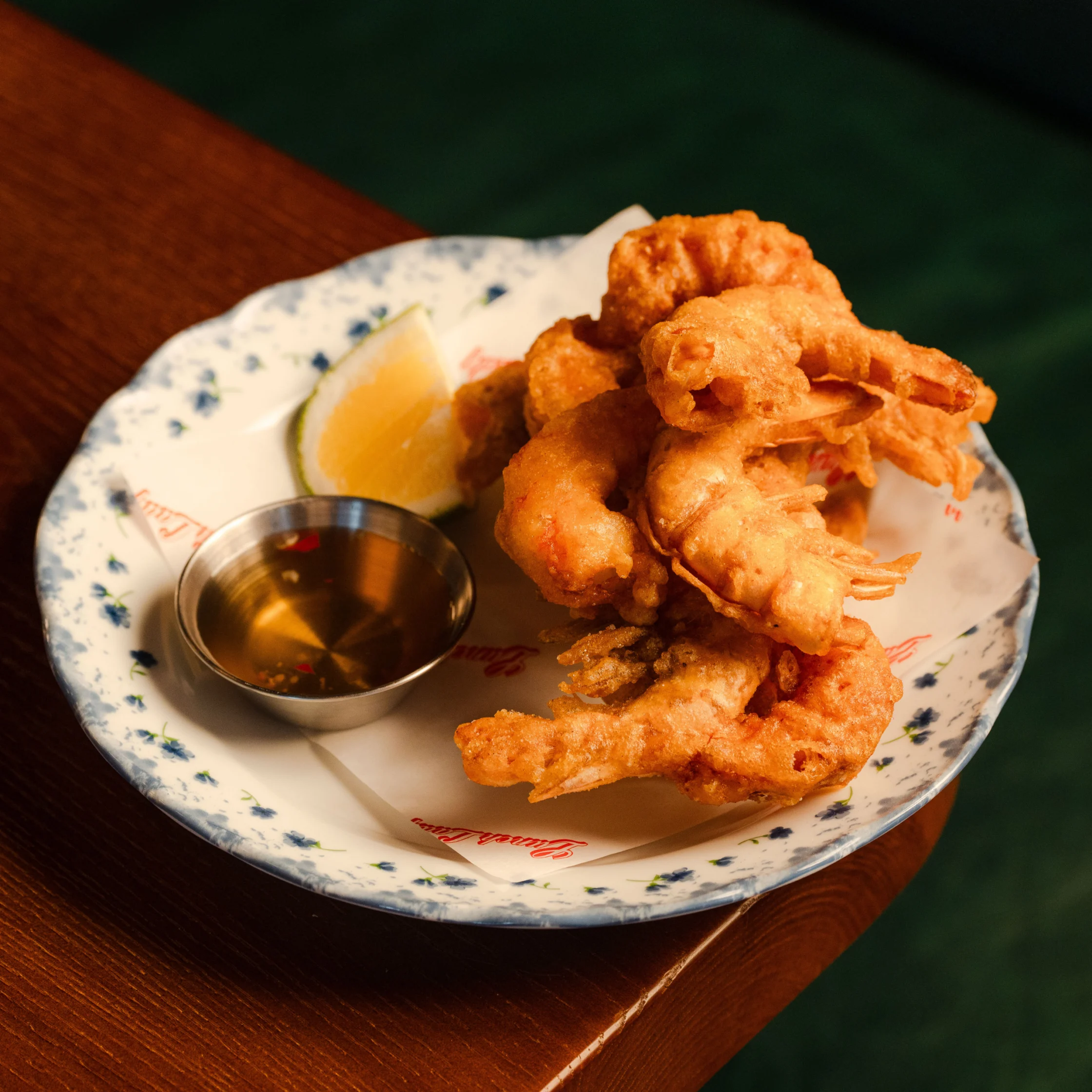 Golden, crispy fried shrimp piled on a blue-and-white plate with a lemon wedge and dipping sauce; a warm, appetizing restaurant scene.