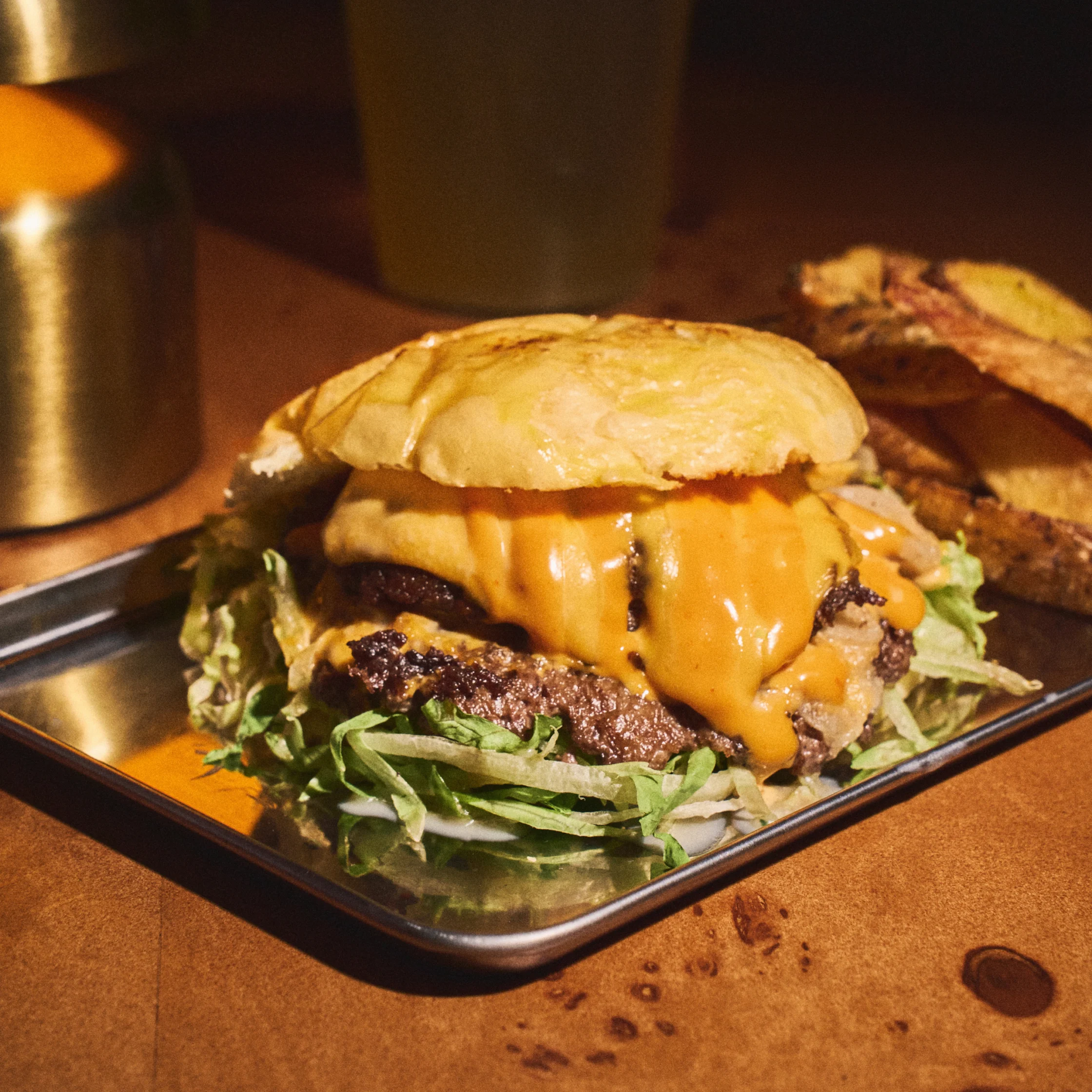 A towering cheeseburger on a metal tray, piled with grilled beef, melted orange cheese, lettuce, and a toasted bun in warm, inviting light.