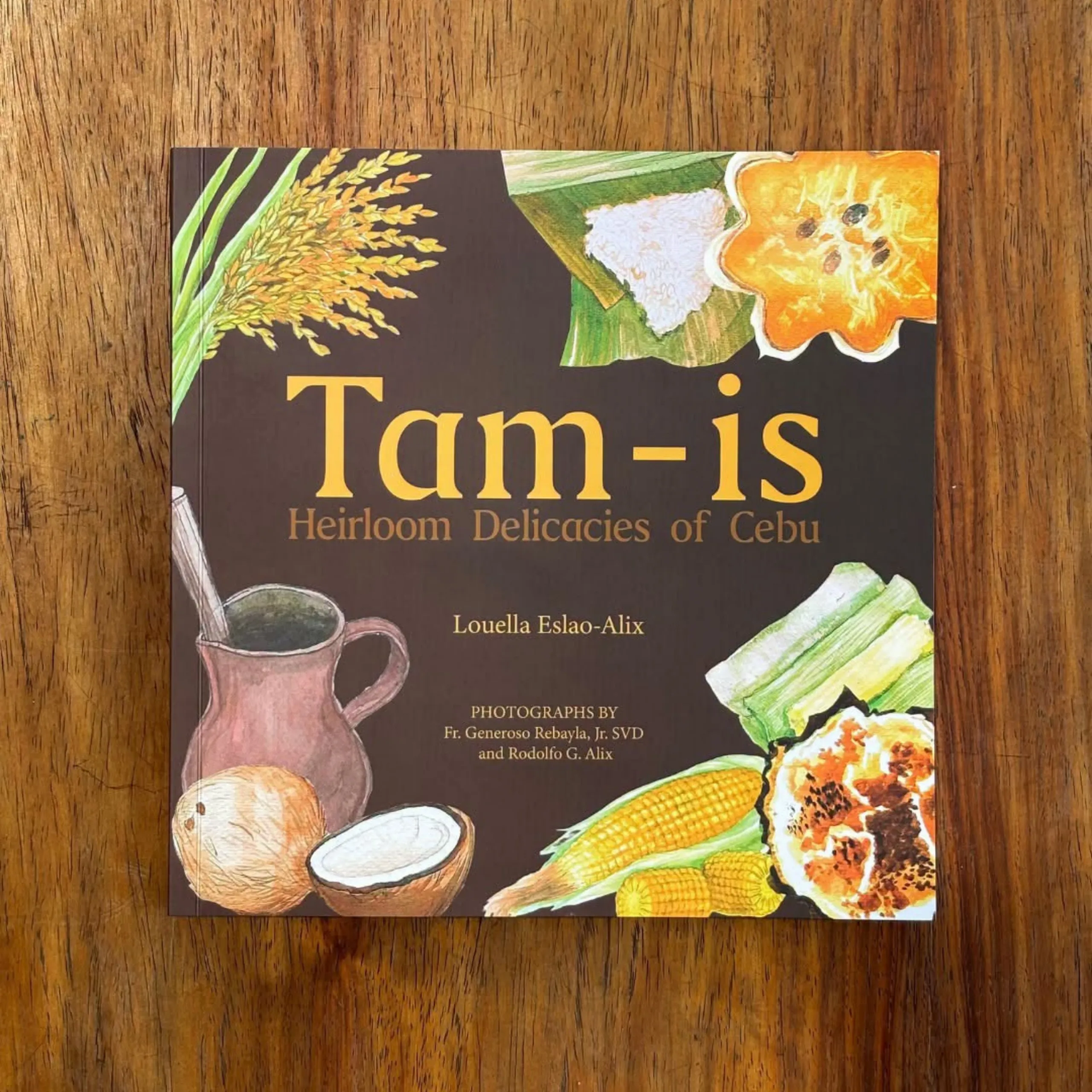 Cookbook titled “Tam-is: Heirloom Delicacies of Cebu” on a wooden table, illustrated with local grains, corn, coconut, and prepared dishes; warm, inviting tone.