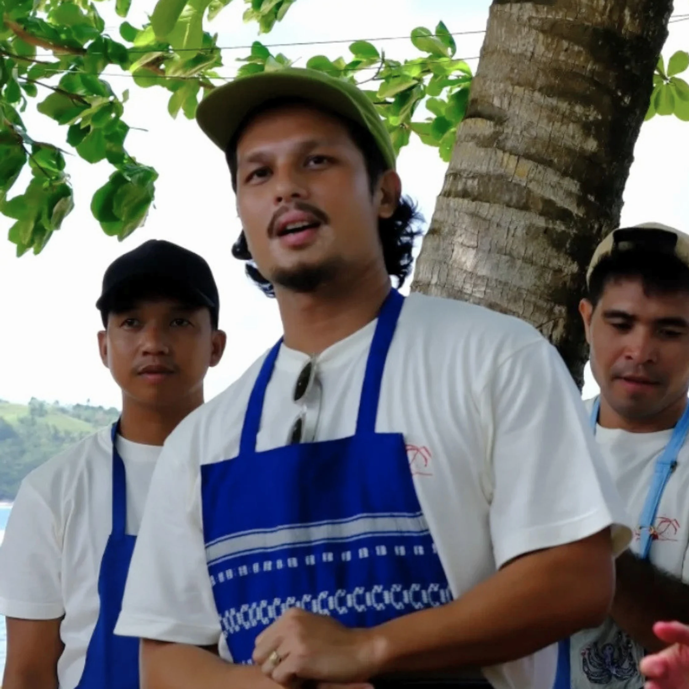 Three men in white shirts and blue aprons stand beneath a leafy tree; the central man in a green cap speaks, creating a casual, attentive outdoor scene.
