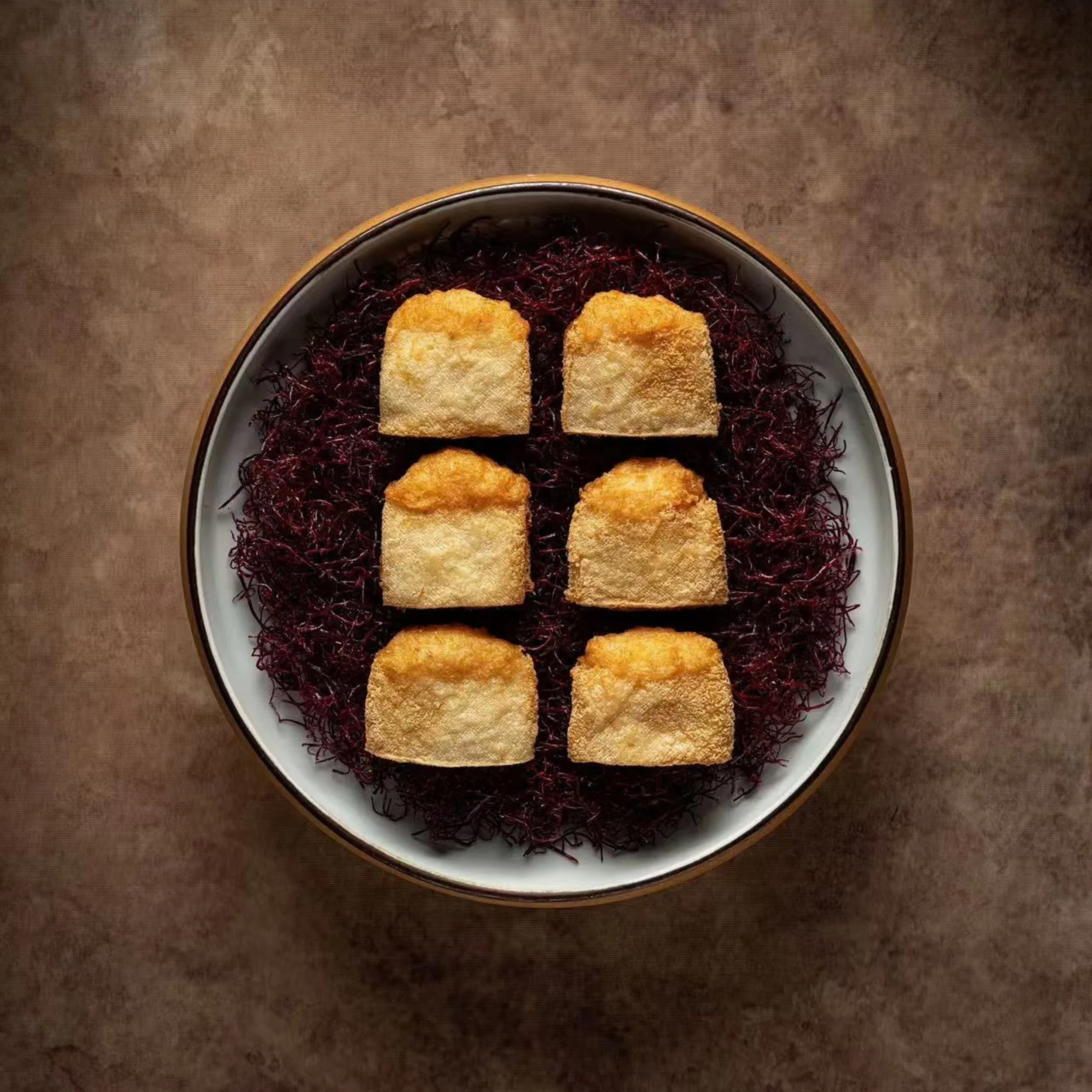 Overhead view of six golden, rectangular pastries arranged in two columns on dark shredded filling in a ceramic bowl, creating a rustic, inviting food presentation.